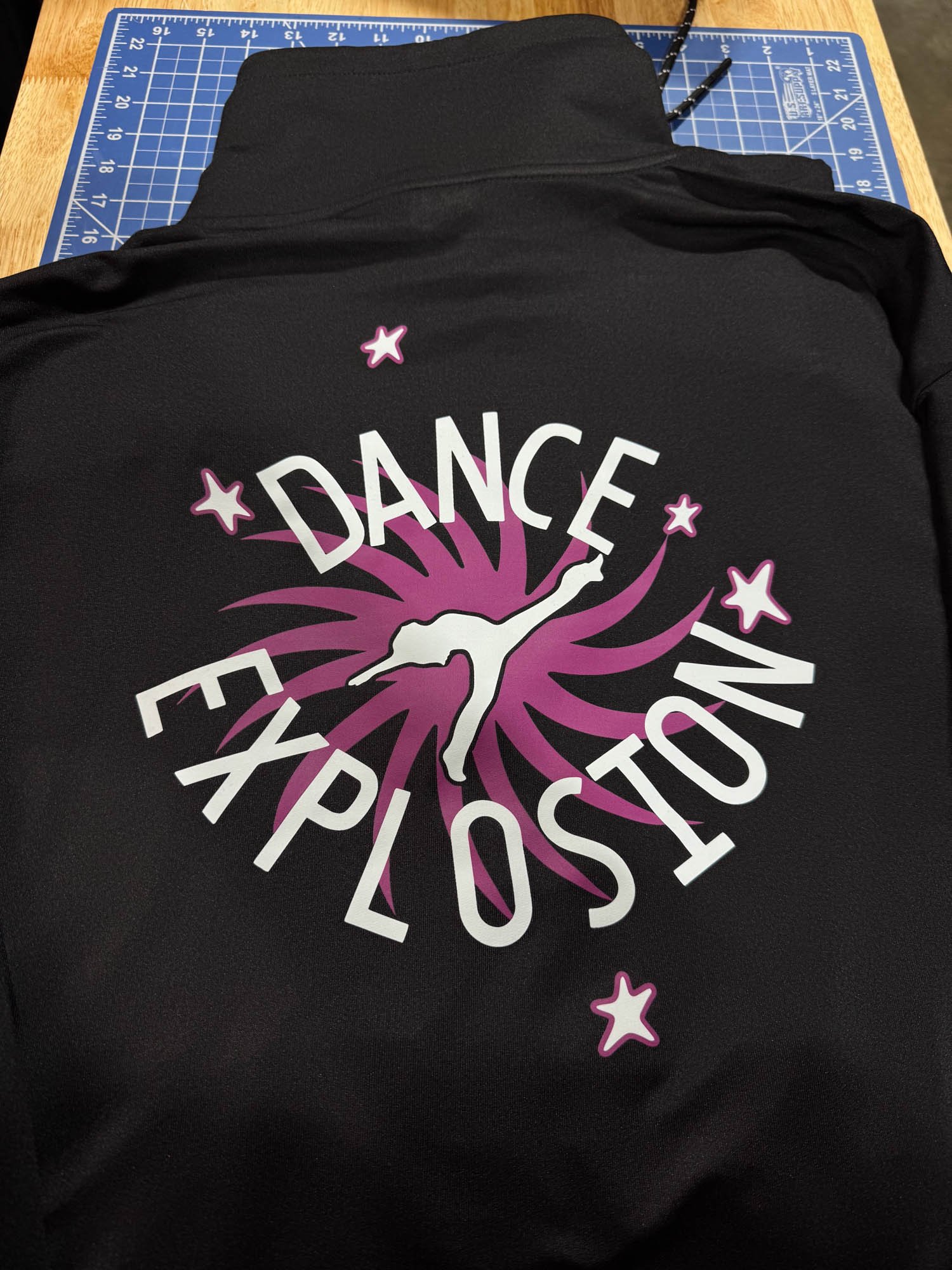 A black garment with a logo that says 'Dance Explosion' featuring a pink spiral burst and a white silhouette of a dancer in an arabesque pose, surrounded by five pink-outlined stars, placed on a wooden table with a blue cutting mat nearby.