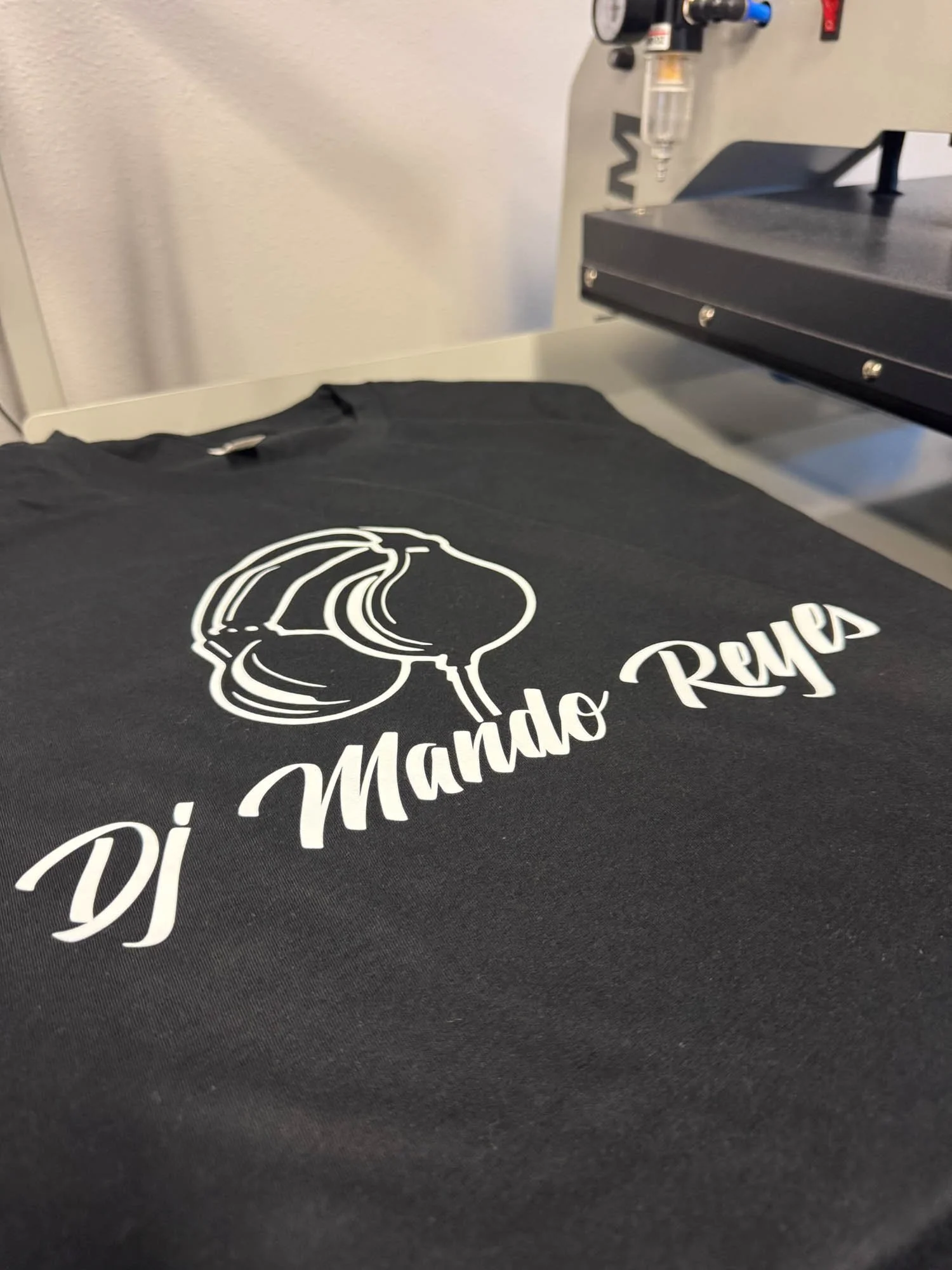 Black t-shirt on a printing machine with a white graphic and text that reads 'DJ Mando Reyes'.