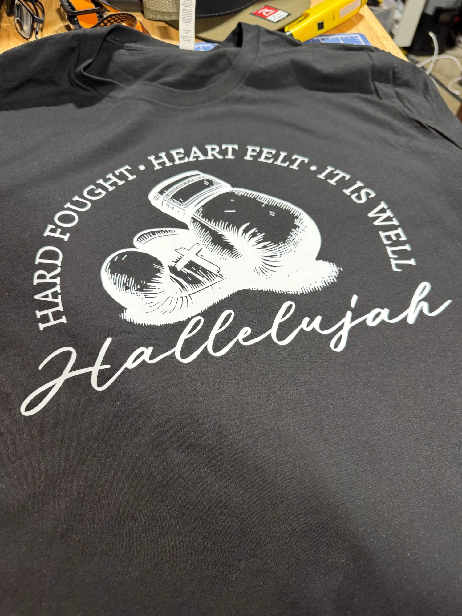 Black T-shirt with white graphic of a nail and the text "Hard fought • Heart felt • It is a well Hallelujah" displayed on a flat surface.