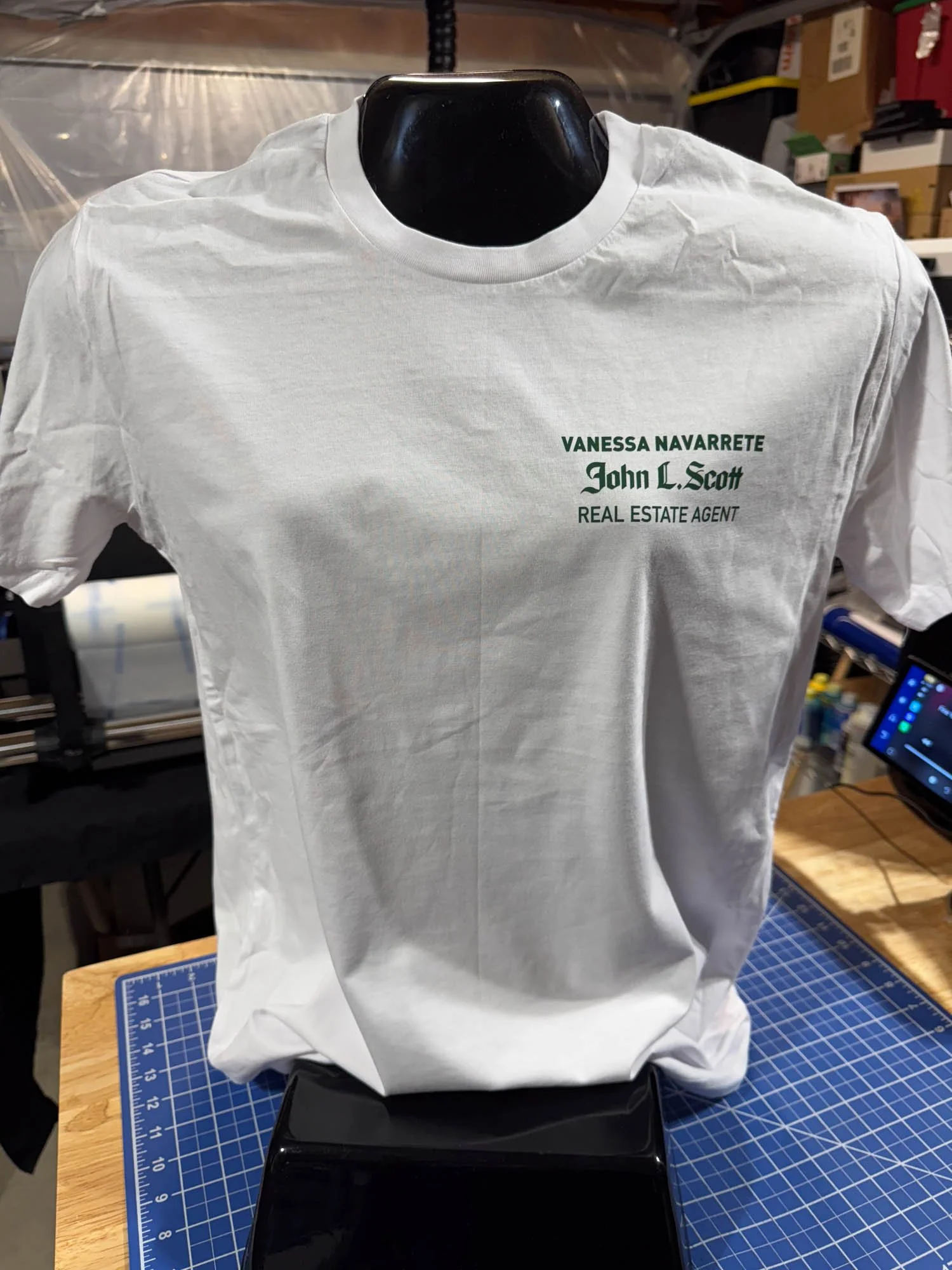 White T-shirt on a display stand with printed green and black text that reads: 'Vanessa Navarrete, John L. Scott, Real Estate Agent'.