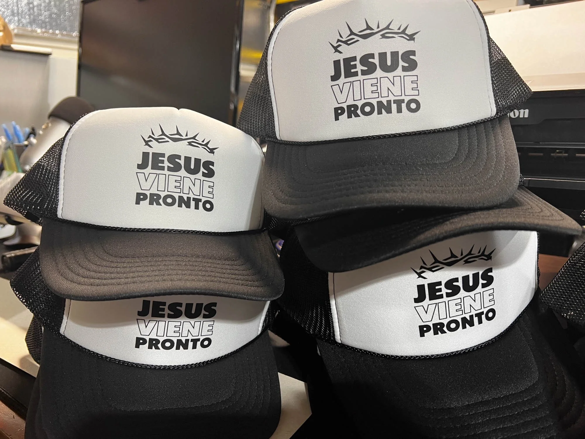 A collection of black and white trucker hats with a phrase in Spanish that translates to 'Jesus is Coming Soon' printed on the front. Some hats are stacked, showing both the front and side views.