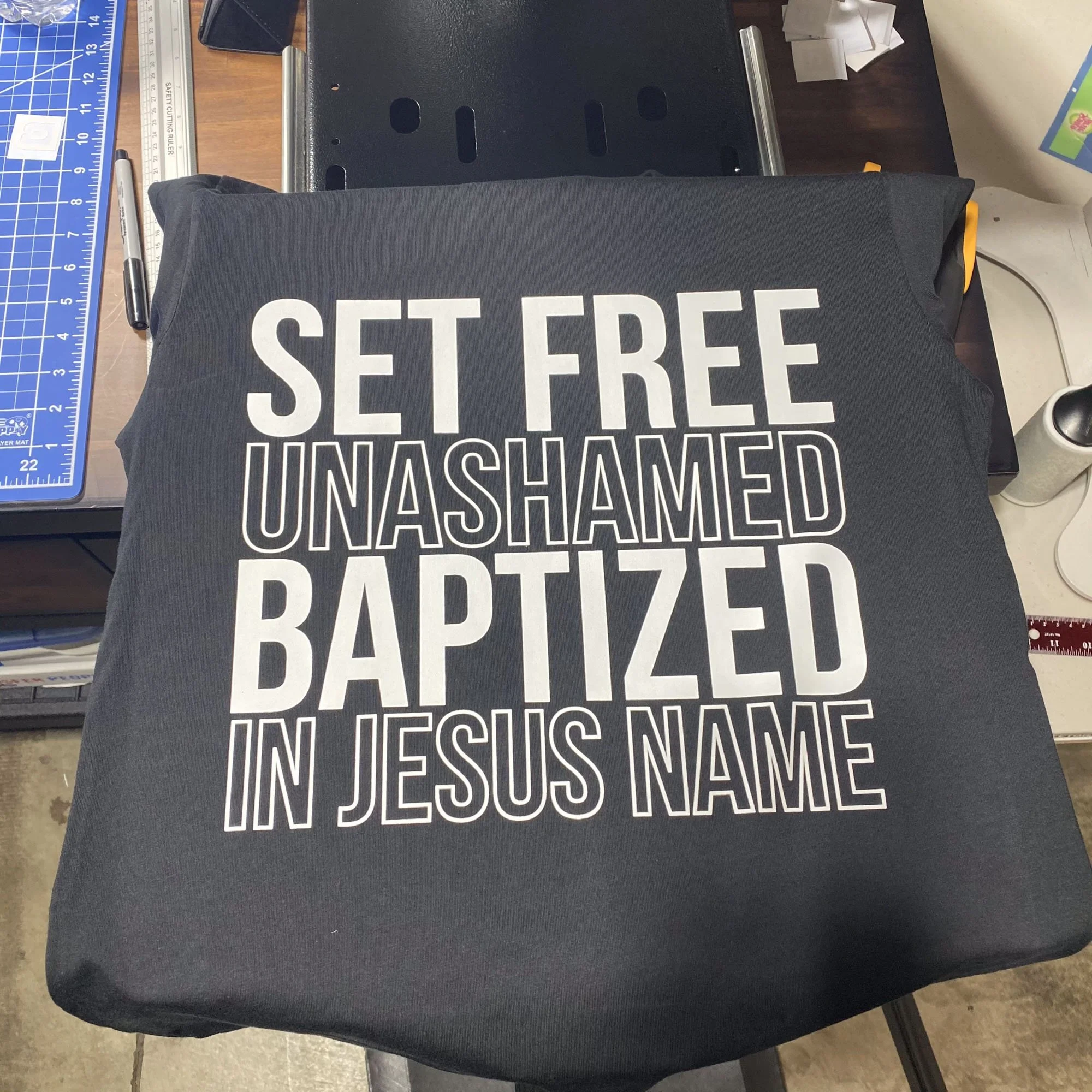 A black pillow with the words 'SET FREE UNASHAMED BAPTIZED IN JESUS NAME' printed in white letters, placed on a desk.