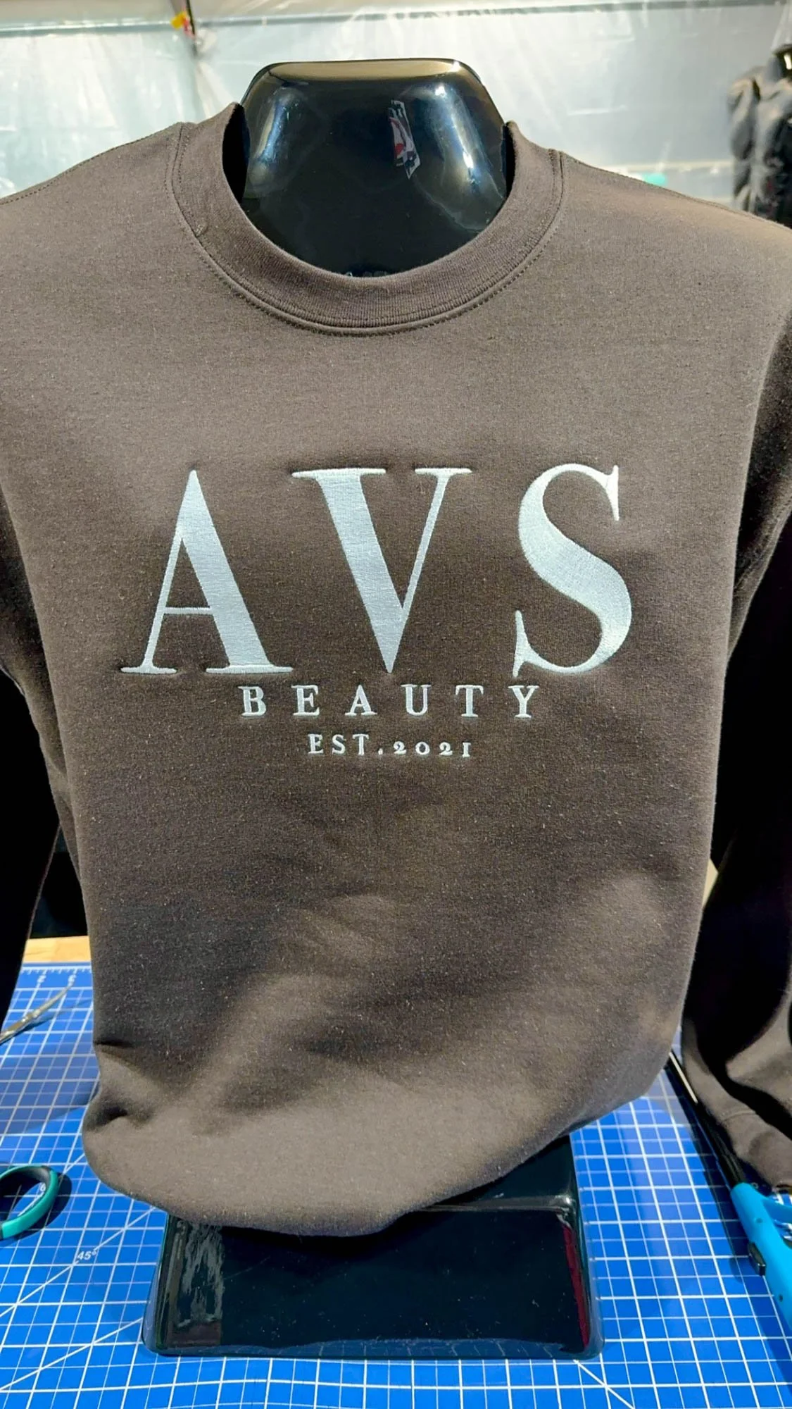 A brown sweatshirt displayed on a black mannequin bust. The sweatshirt has white embroidered text that says "AVS BEAUTY EST. 2021".