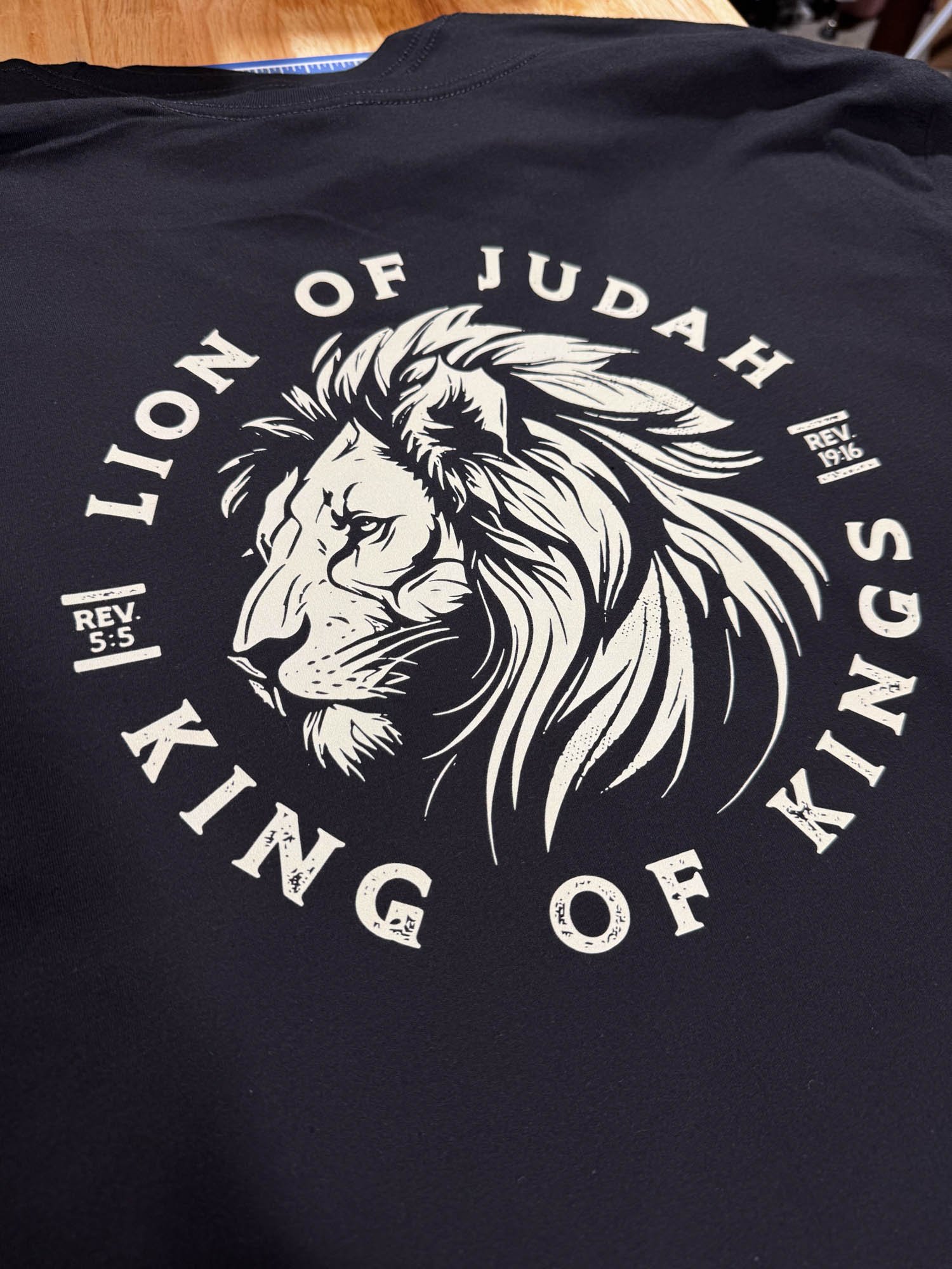 Close-up of a dark t-shirt with a white graphic featuring a lion's head and the text 'Lion of Judah', 'King of Kings', and biblical references Rev. 5:5 and Rev. 19:16.