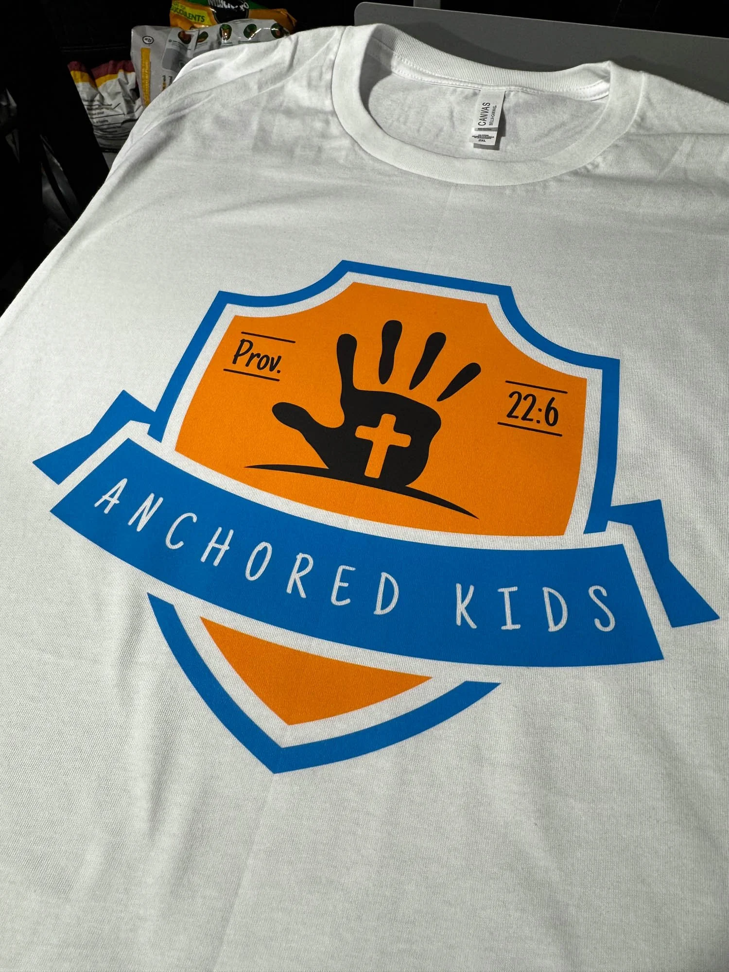 A white T-shirt with a logo design that includes an orange shield with a black hand making a 'stop' gesture, a gold cross on the wrist, and the text 'Prov. 22:6' and 'Anchored Kids' on a blue banner.