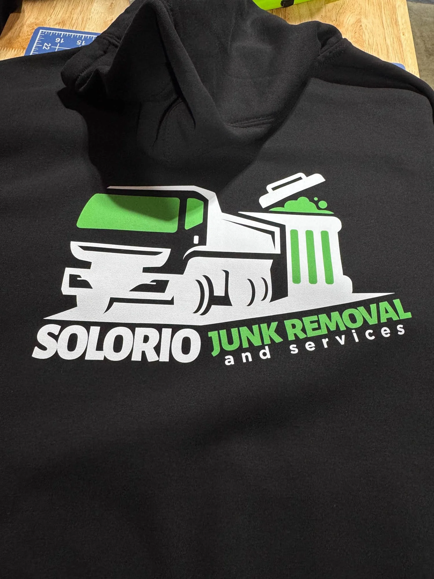 Black hoodie with a logo for Solorio Junk Removal, featuring a graphic of a truck and trash can in green and white, and the company name and services in bold text.