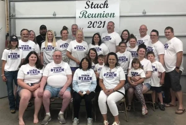 Group of people attending the Stach Reunion 2021, with some wearing matching T-shirts that say 'Team Stach', gathered in a large room with a banner above that reads 'Stach Reunion 2021'.
