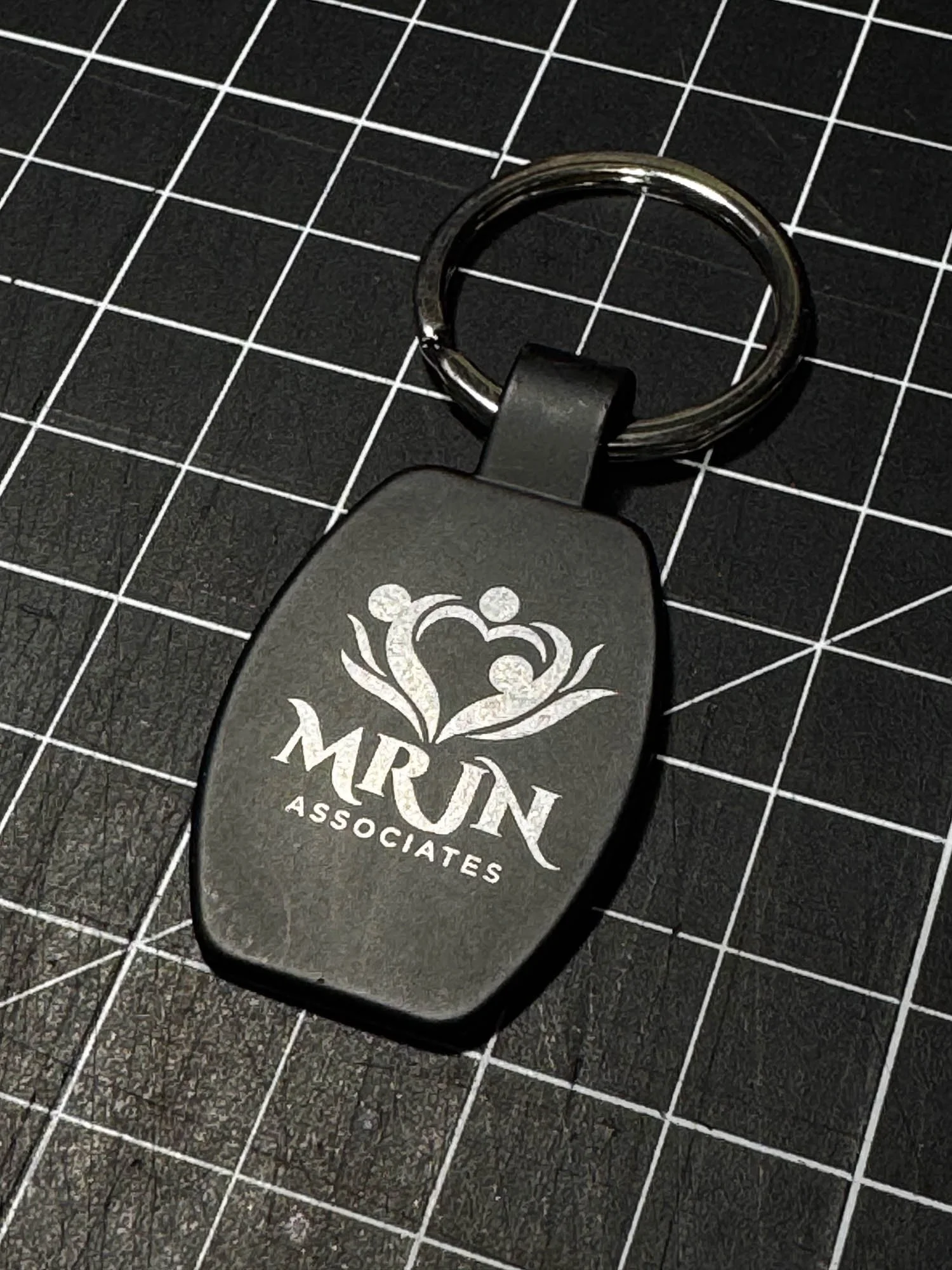 A black keychain with a metal ring attached, displaying the logo and text for 'MRUN ASSOCIATES' on a black grid cutting mat.