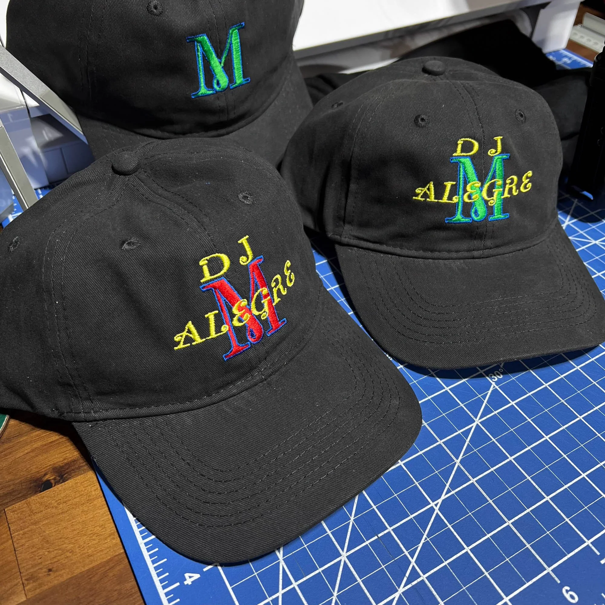 Black caps with colorful embroidered text that reads "DJ ALLEGRE" and initials "M" on the front, placed on a blue cutting mat.