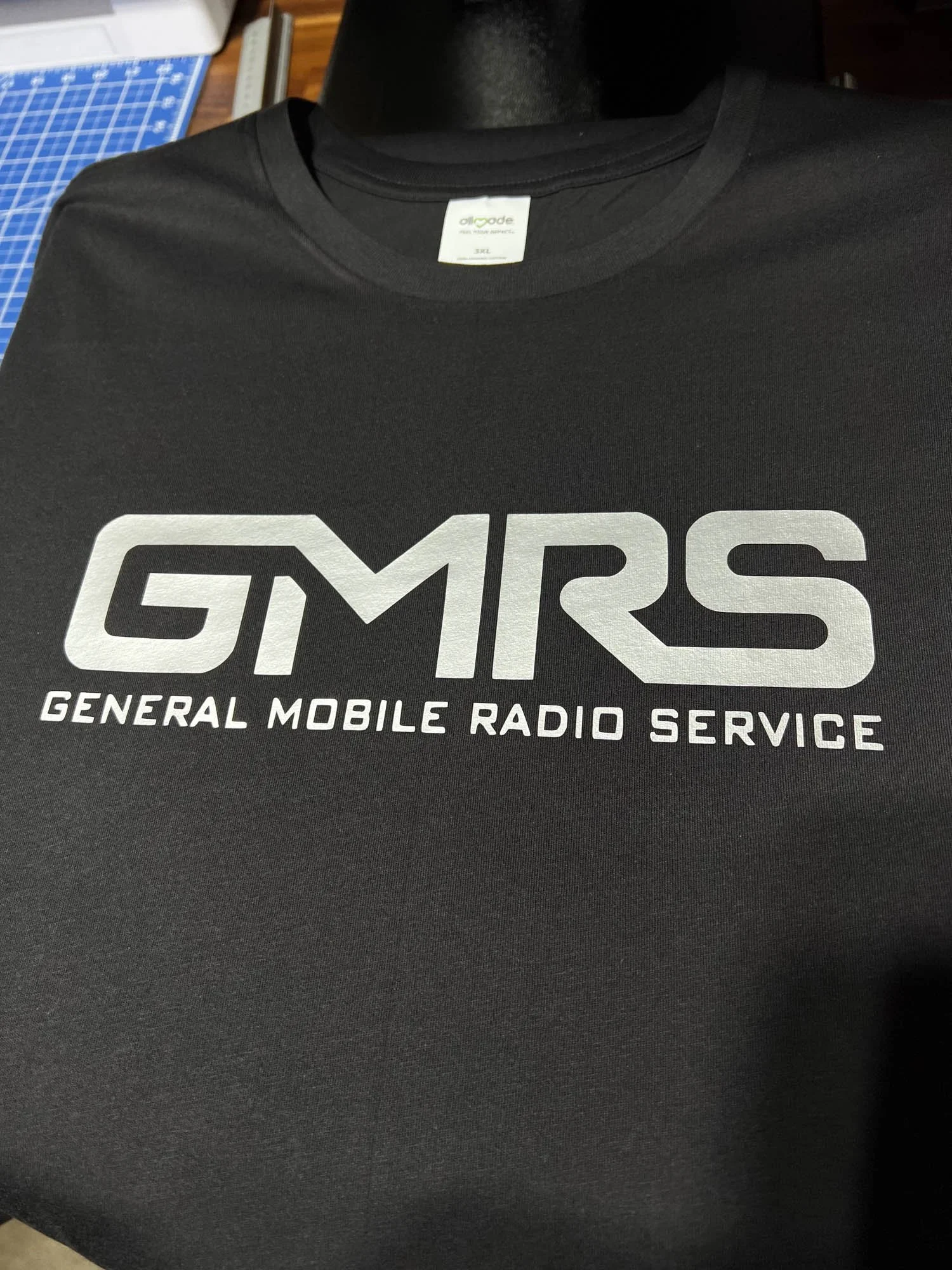 Black T-shirt with white text reading GMRS and General Mobile Radio Service laid flat on a surface, with a cutting mat and other tools in the background.