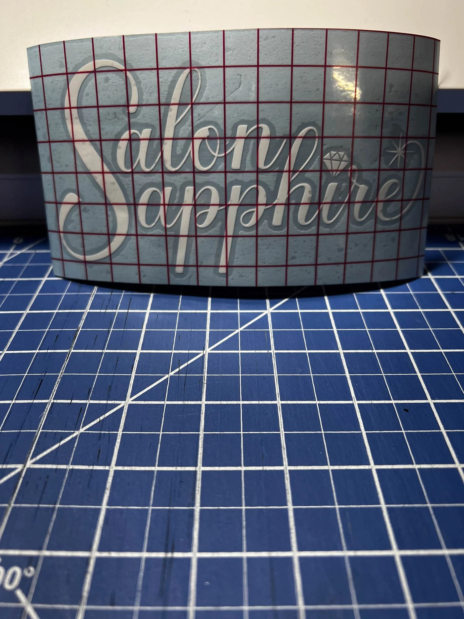 A curved sign with the text "Salon Sapphire" in cursive letters, placed on a blue cutting mat with grid lines.
