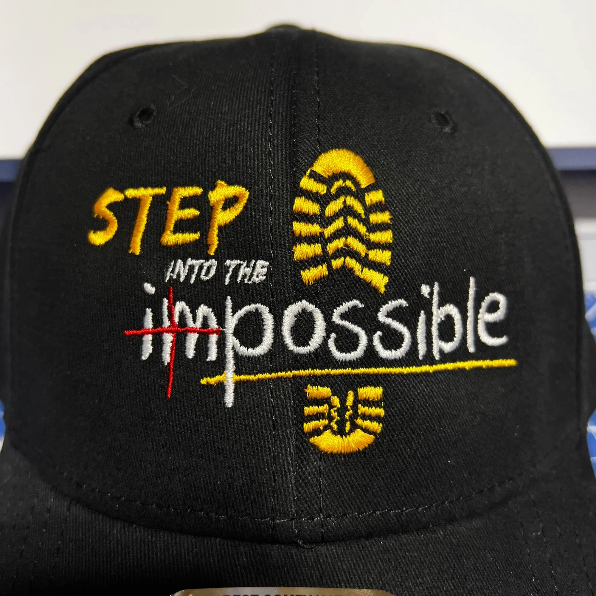 Black baseball cap with yellow, white, red embroidery reading 'STEP INTO THE impossible' with a yellow and black footprint design.