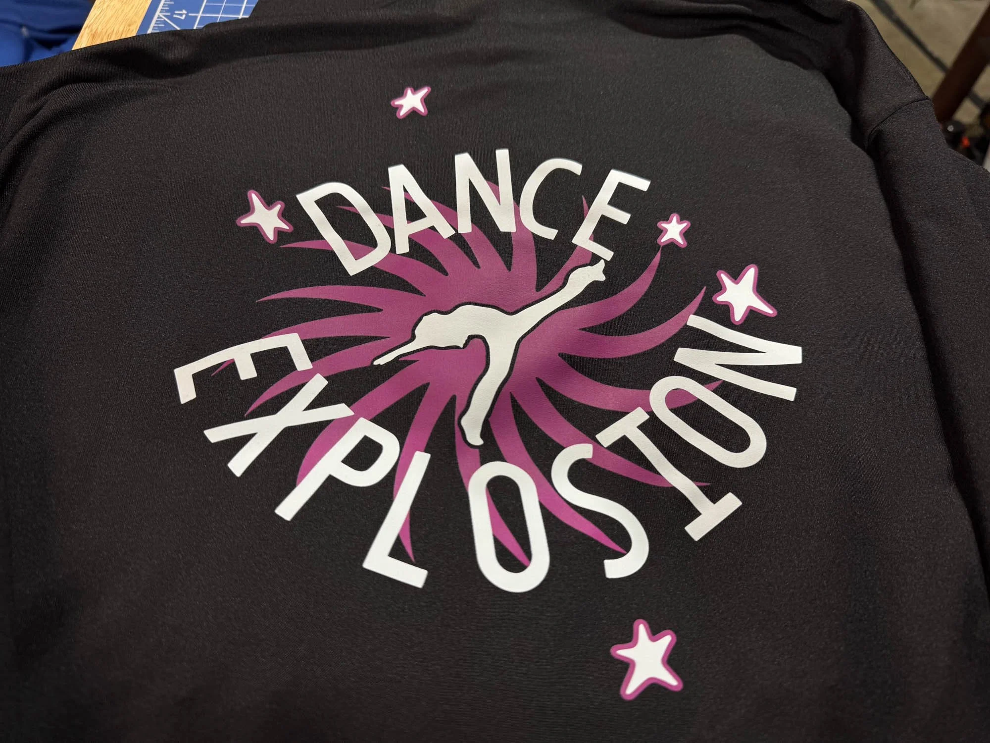 Black fabric with a colorful design featuring a female figure in a dance pose, surrounded by pink and purple star shapes and swirling lines, with the words 'DANCE EXPLOSION' in bold white letters.
