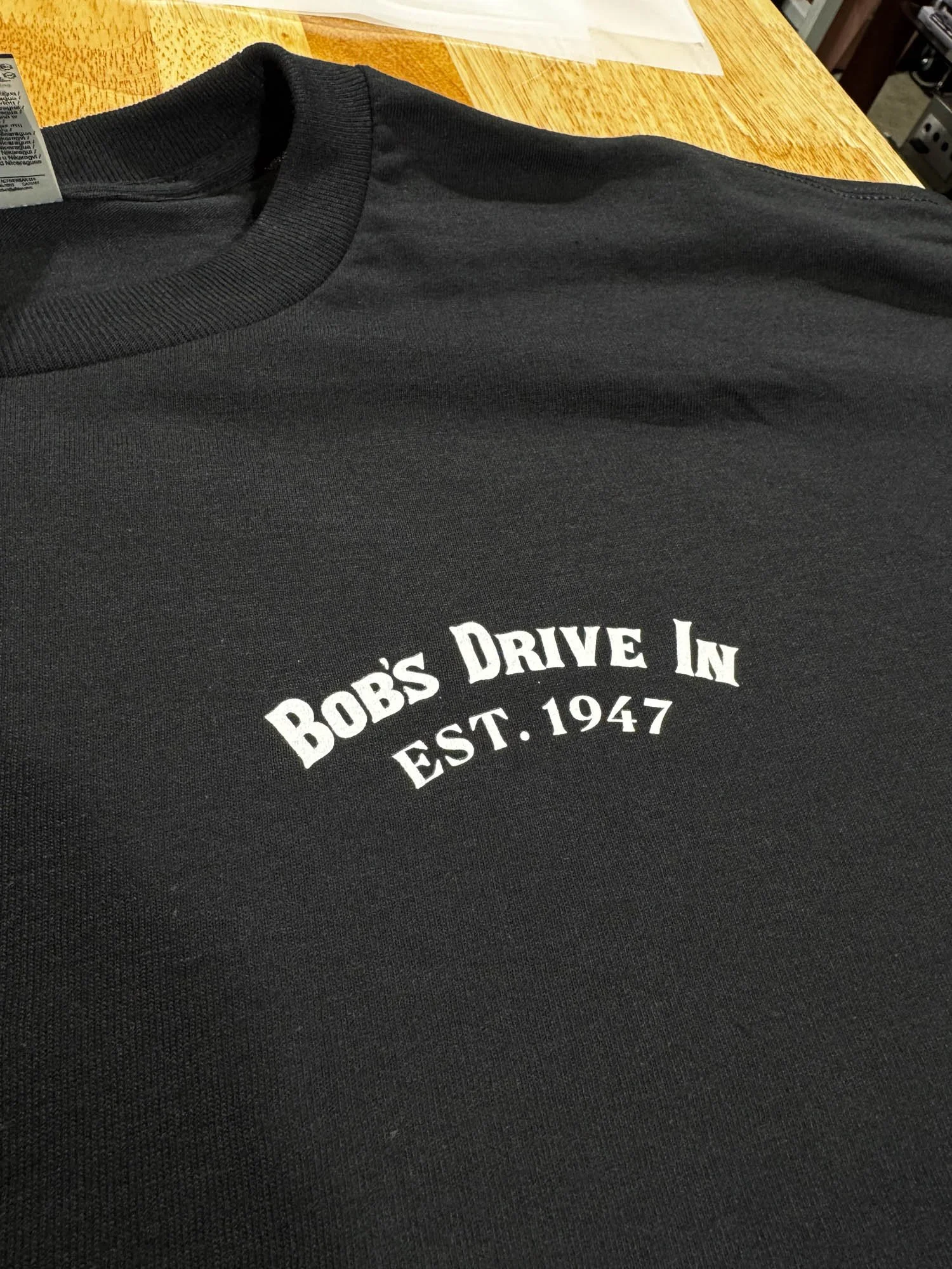 Close-up of a black sweatshirt with white text reading 'Bob's Drive-In EST. 1947' resting on a wooden surface.