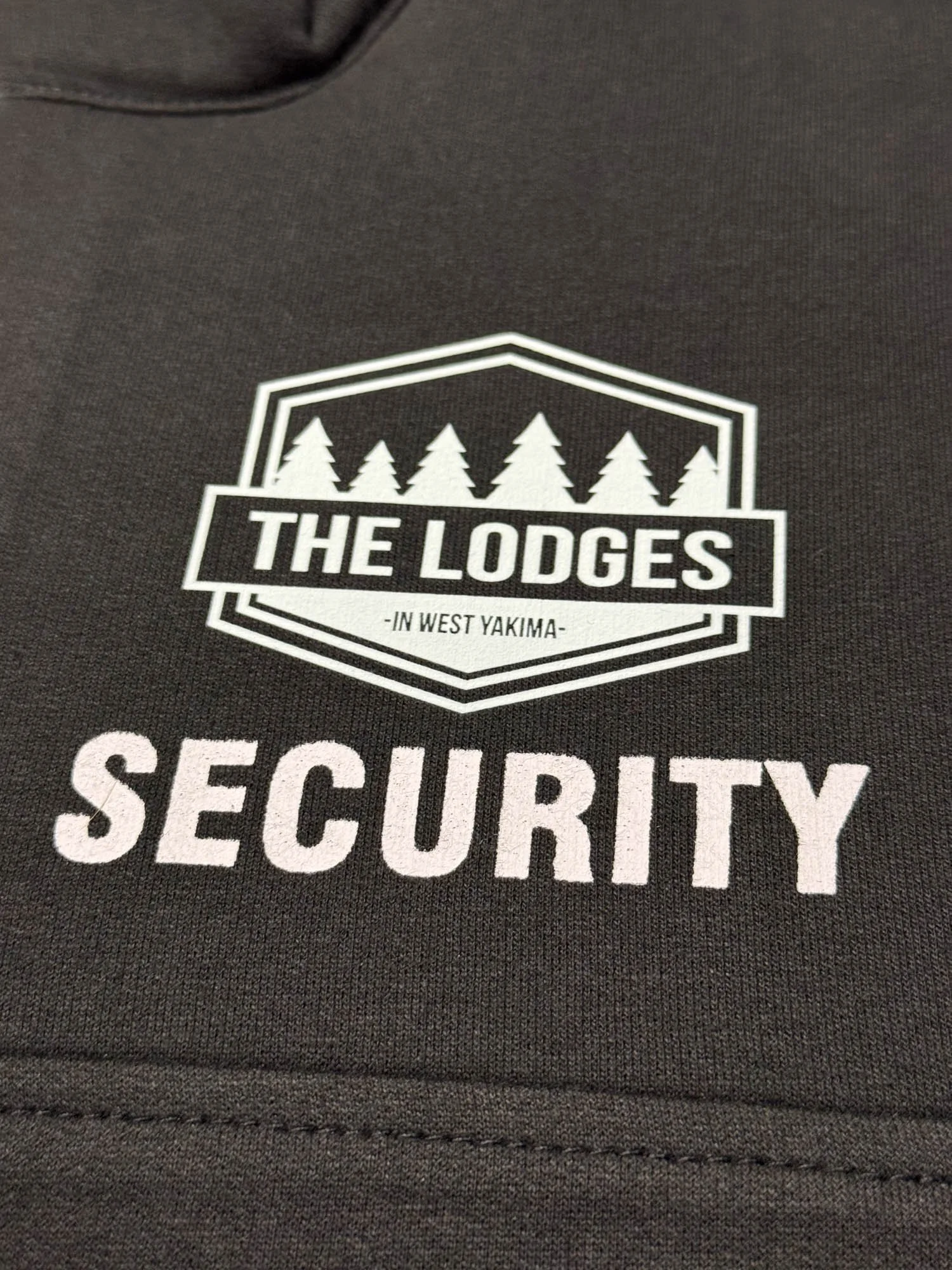 Close-up of a dark grey uniform shirt with a logo that says "The Lodges in West Yakima" and "Security" underneath, featuring a pine tree design.