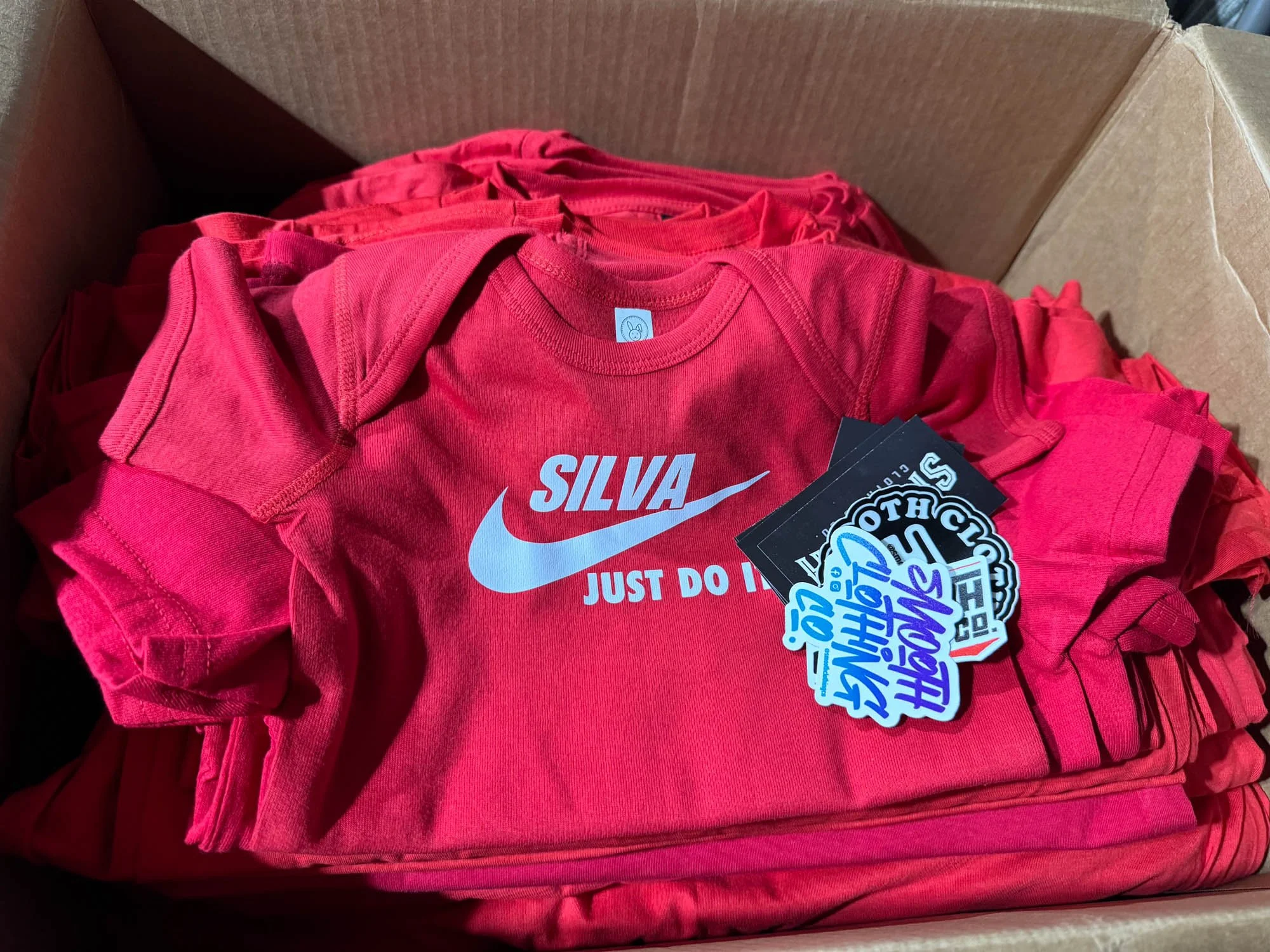 A cardboard box filled with multiple red Nike T-shirts, one of which is prominently displayed with the name 'Silva' and the Nike logo on the front, along with tags attached to the shirts.
