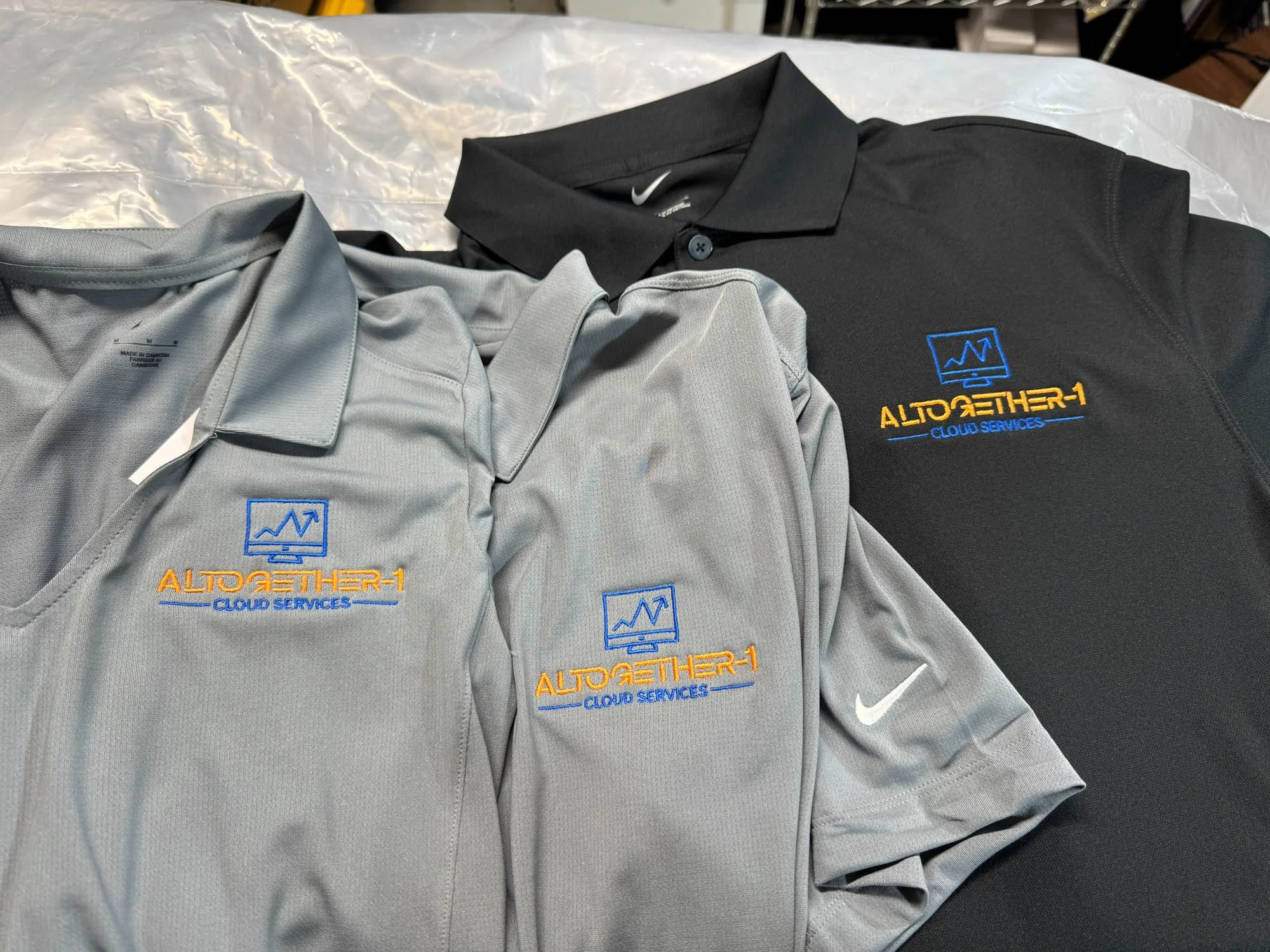 Three polo shirts with company logo embroidered on the left chest, two in gray and one black, laid out on a plastic surface. The logo features a blue monitor with a graph line and the text 'ALTOETHER-1 CLOUD SERVICES' in orange and blue.