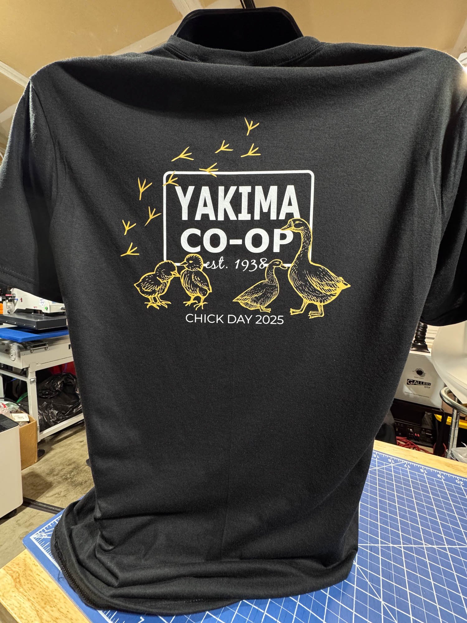 Black T-shirt with yellow and white printed design featuring ducks, the words "YAKIMA CO-OP," establishment date 1938, and the message "CHICK DAY 2025," hanging on a mannequin in a room with a cutting mat and various objects in the background.