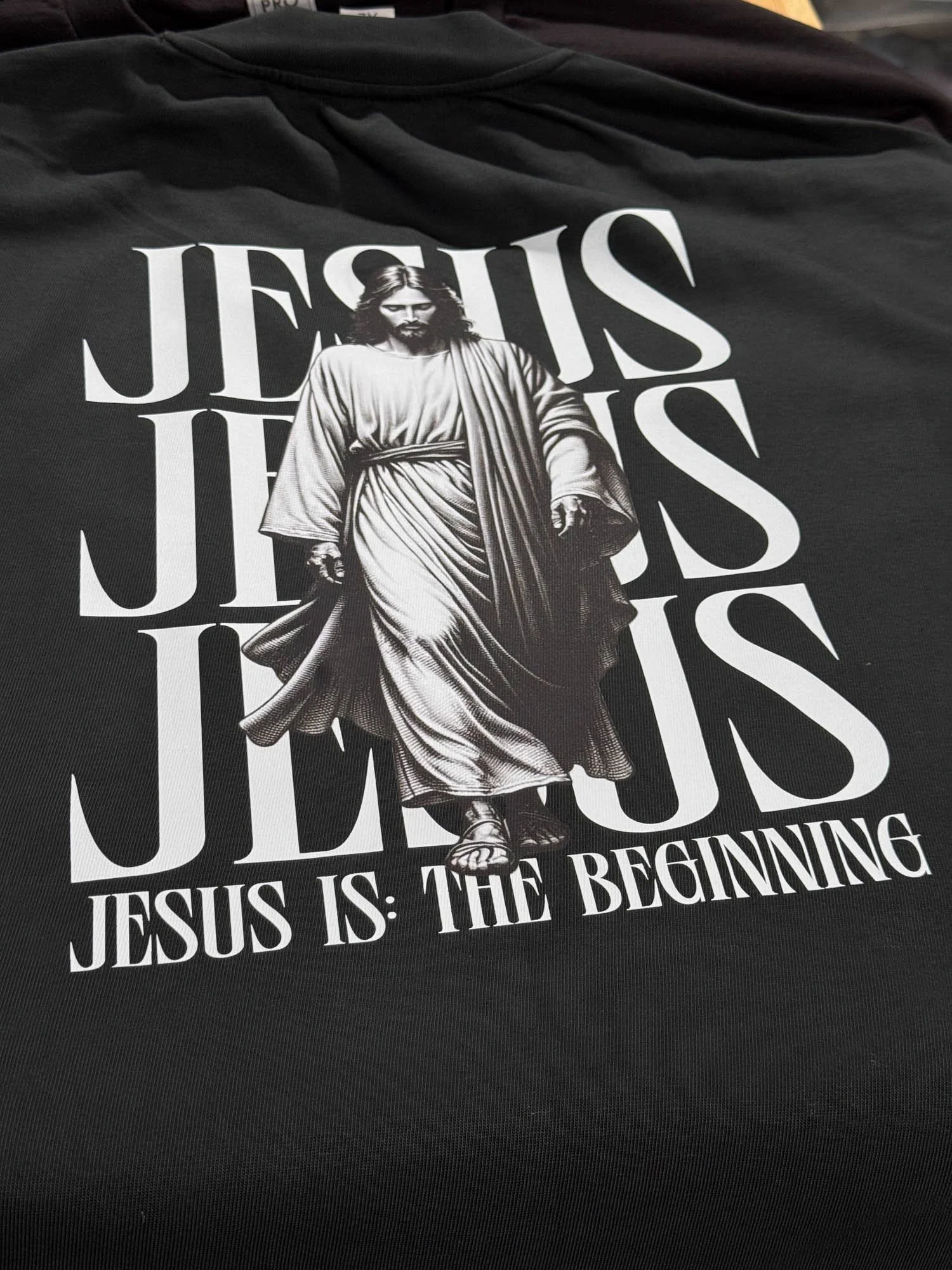 Black T-shirt with white print of Jesus Christ walking, overlaid with large text reading "JESUS IS" and smaller text "JESUS IS: THE BEGINNING" underneath.