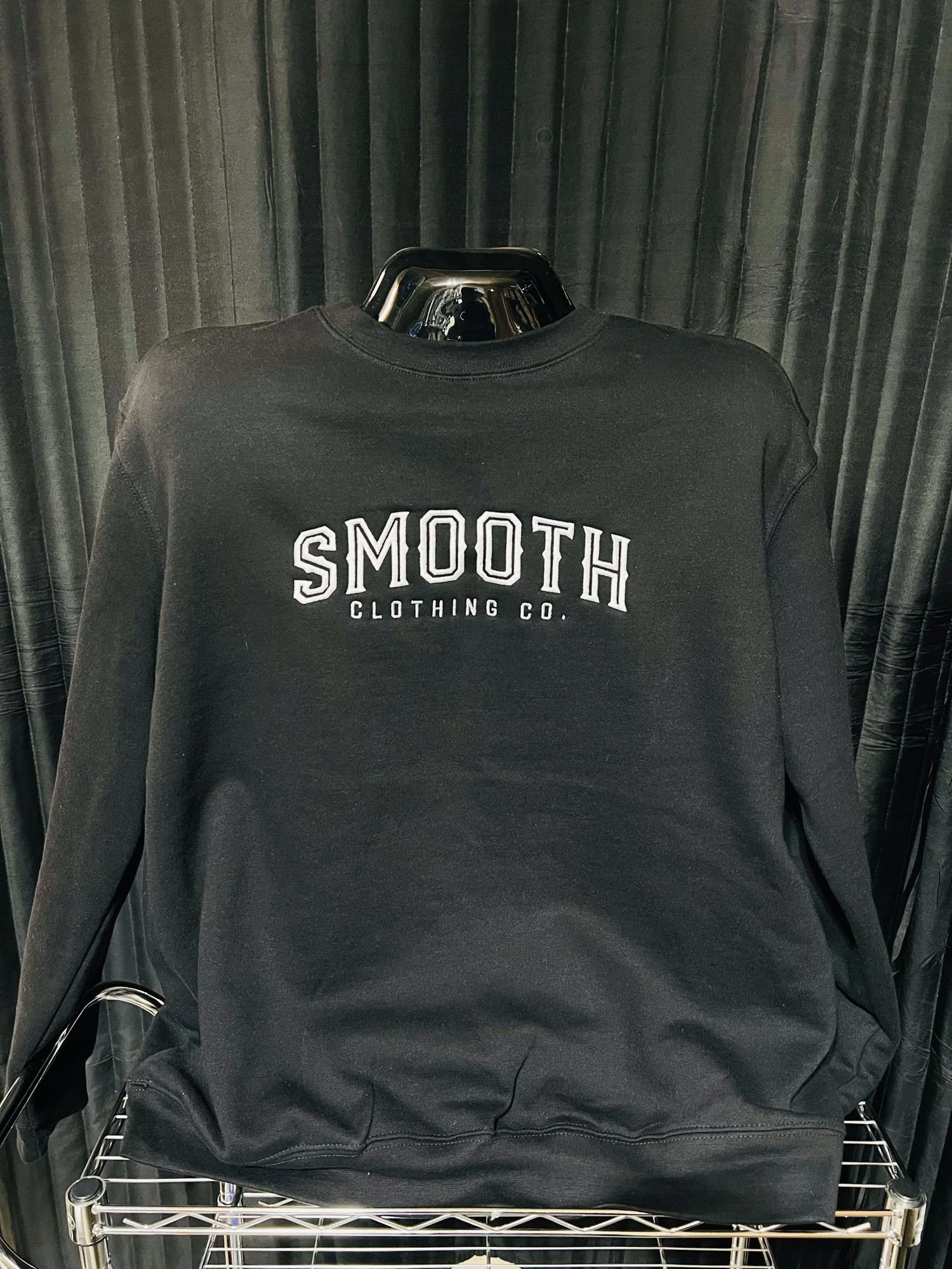 Black sweatshirt with white embroidered text that says 'SMOOTH CLOTHING CO.' displayed on a mannequin against a black curtain background.