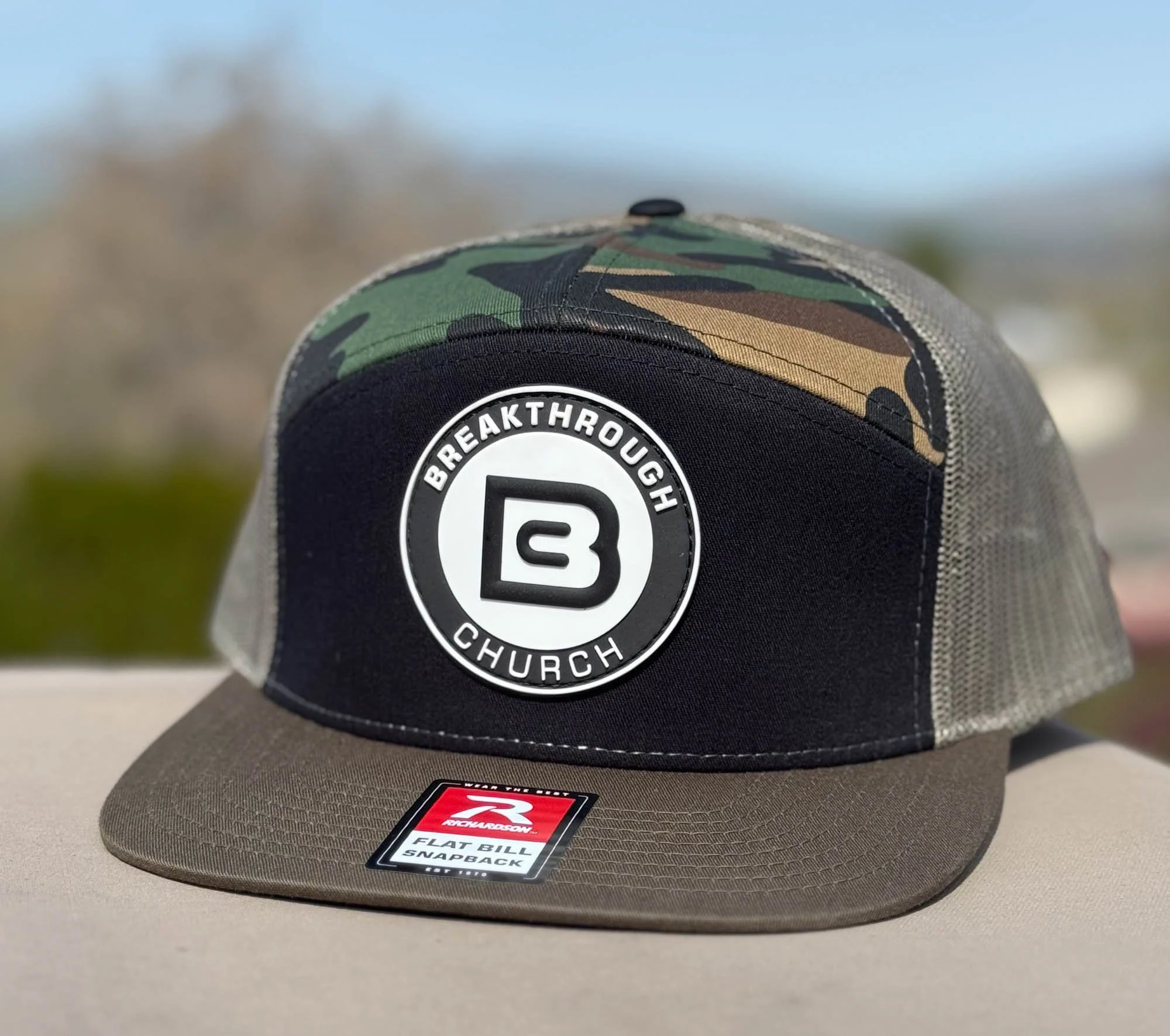 A cap with a camouflage pattern on the crown, a black front panel with a circular patch reading 'Breakthrough Church,' a tan flat bill with a sticker, and gray mesh on the back.