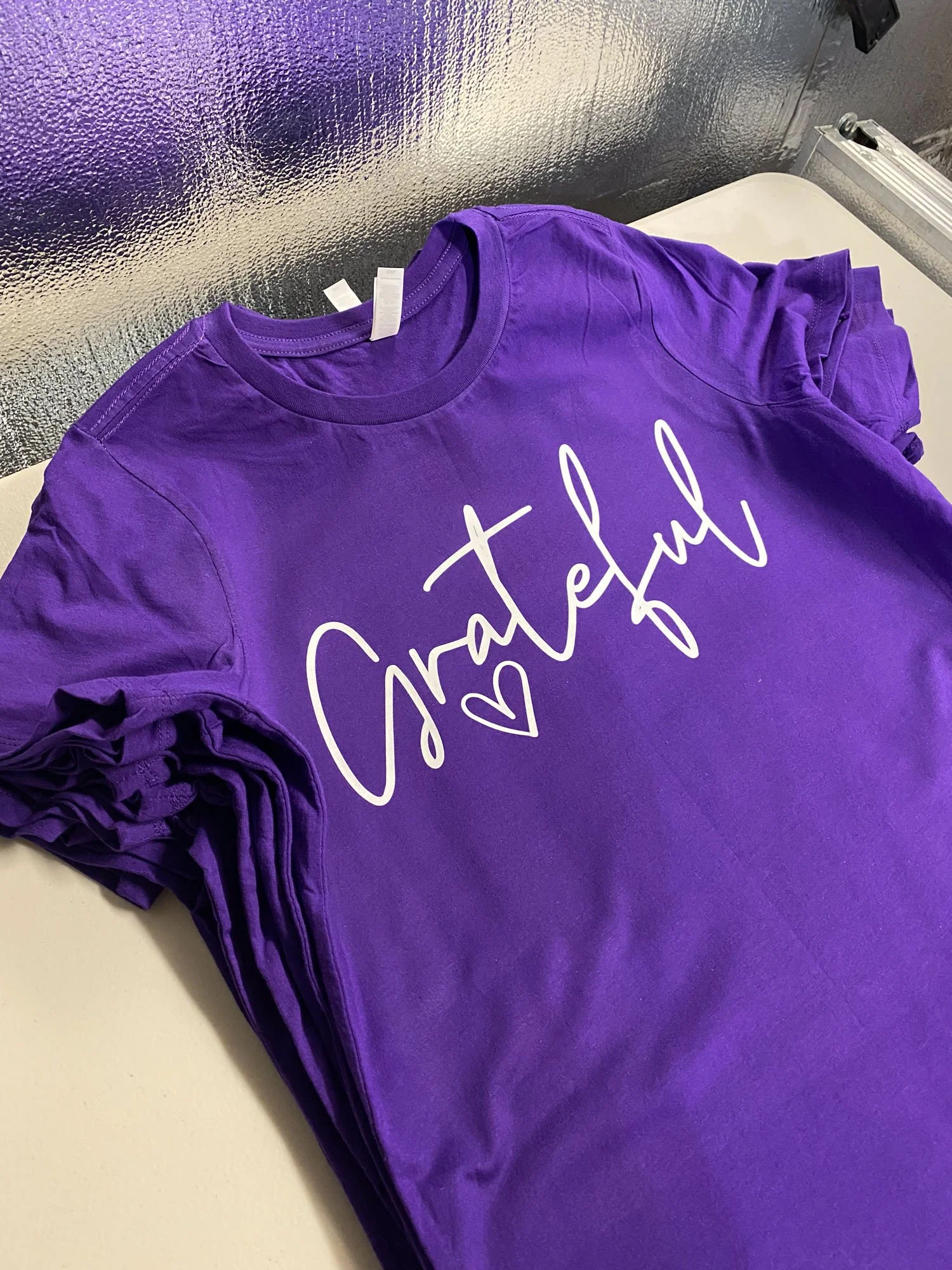 Purple T-shirt with the word 'Grateful' and a heart design printed on it, laid on a white surface.