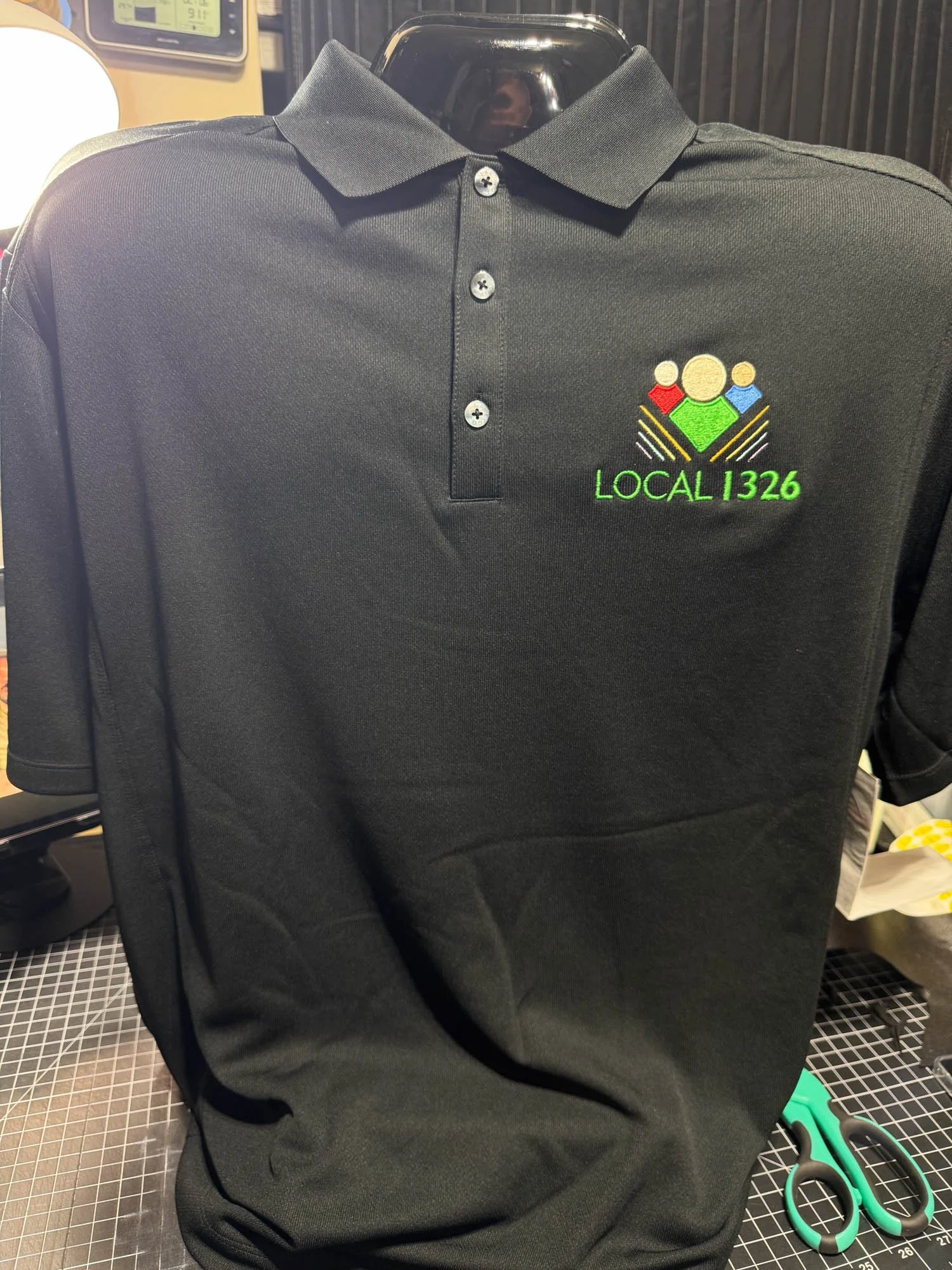 Black polo shirt with a multicolored embroidered logo and green text, displayed on a black mannequin. The logo features three stylized figures and the text reads 'LOCAL 1326'.