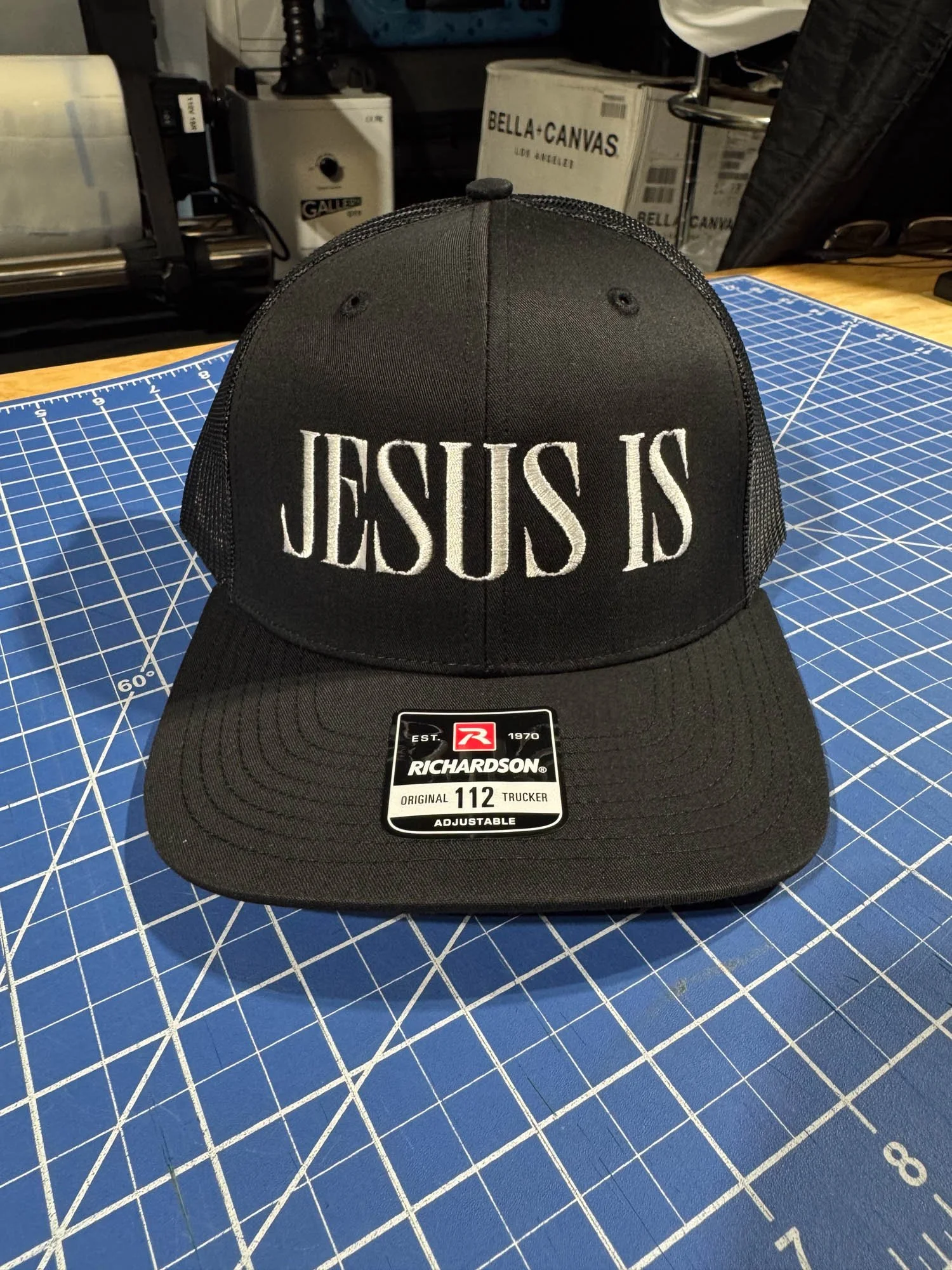 Black Richardson cap with white embroidered text reading 'JESUS IS' on the front, placed on a blue cutting mat with white grid lines.