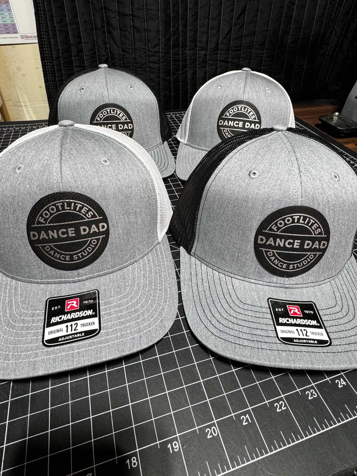 Four gray and black baseball caps with a circular logo that reads 'Footlites Dance Dad' and 'Dance Studio' on the front. The caps are placed on a black cutting mat with grid lines.