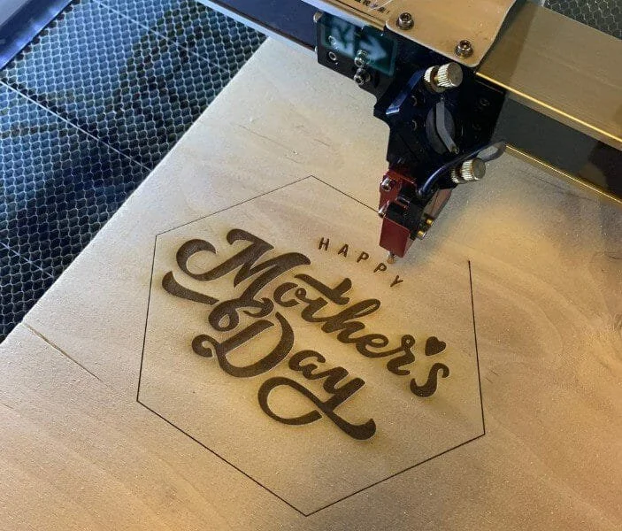 A laser engraving machine is creating a 'Happy Mother's Day' message inside a hexagon on a wooden surface.