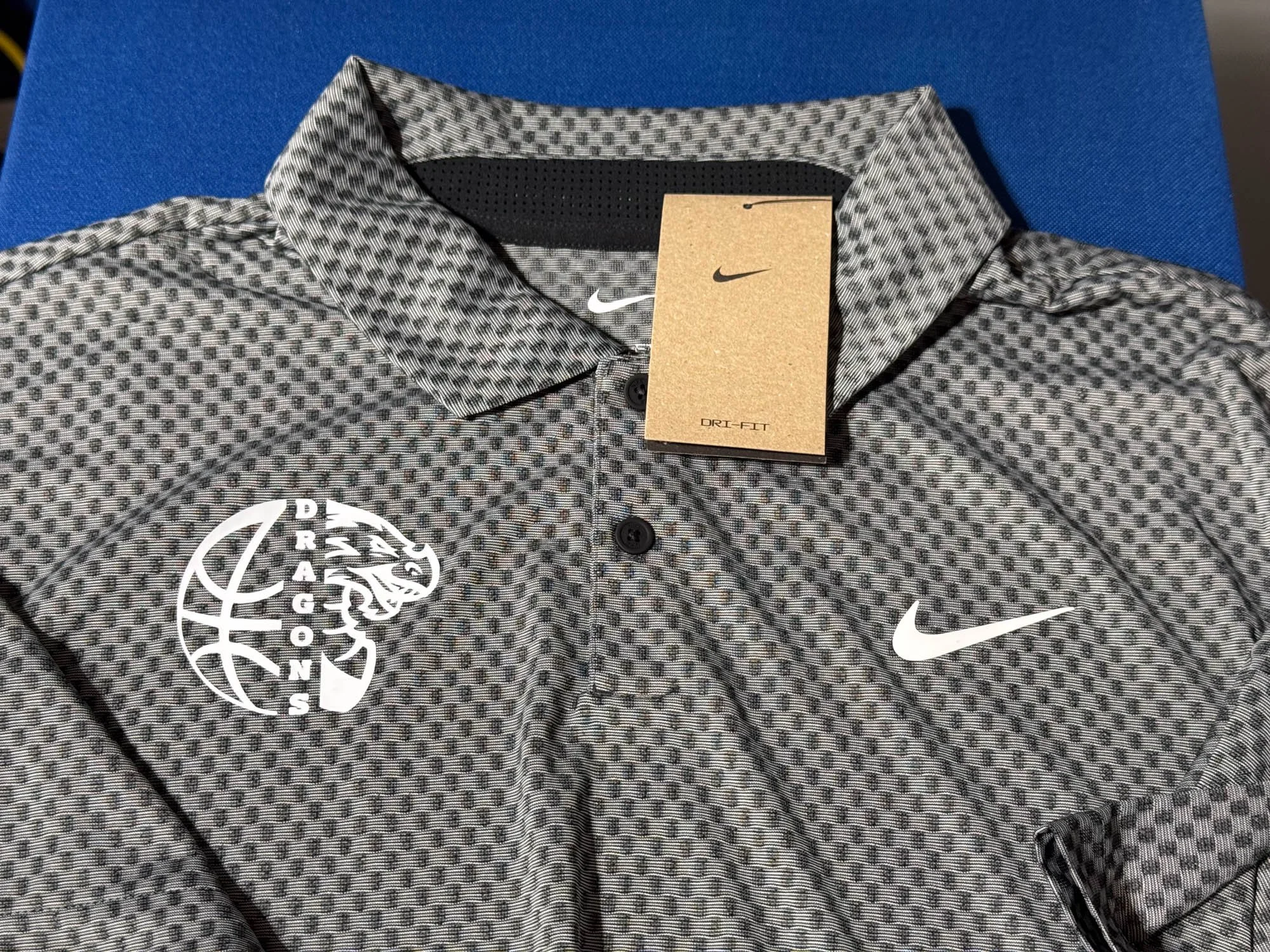 A Nike Sportswear golf polo shirt with a black and white geometric pattern, featuring the Oregon Ducks logo on the left chest and the Nike swoosh on the right chest, laid on a blue surface.
