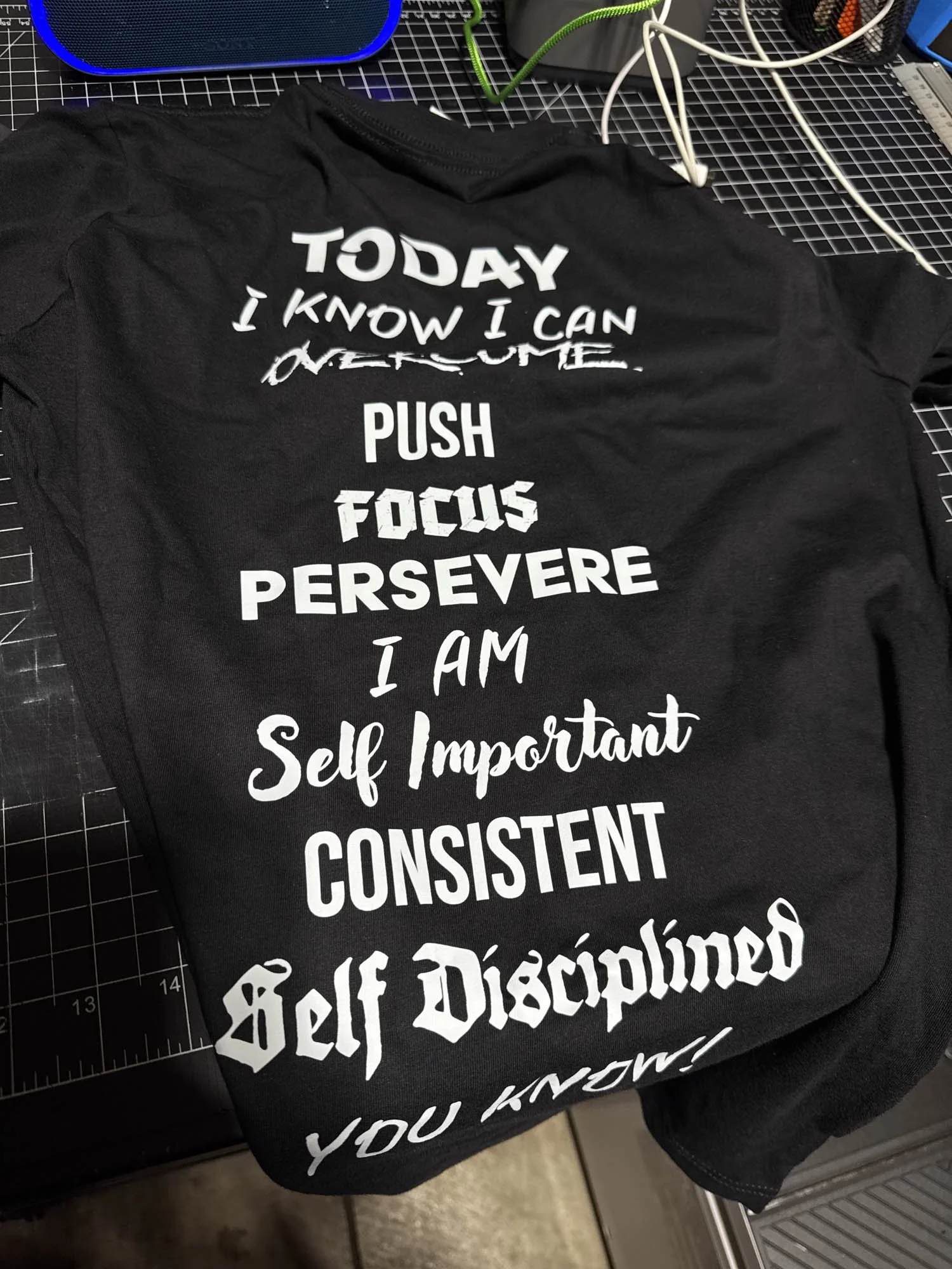 A black T-shirt with motivational text printed in white, featuring words like 'TODAY,' 'PUSH,' 'FOCUS,' 'PERSEVERE,' and phrases encouraging self-importance and discipline, placed on a work surface amidst crafting supplies.
