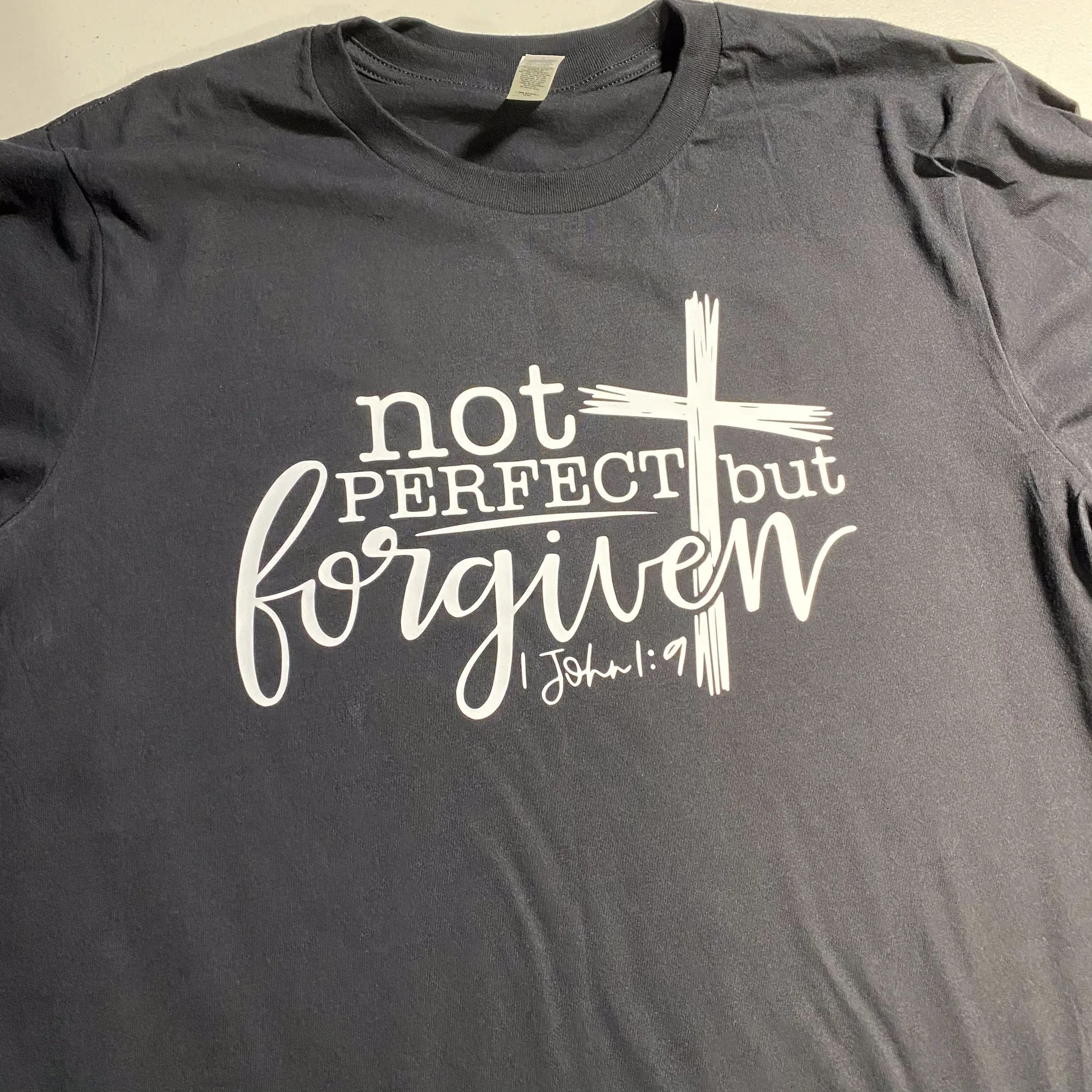 Black T-shirt with white Christian-themed text and design. The text reads, "not perfect but forgiven," with a cross integrated into the design, and the Bible verse reference "John 1:9" below.