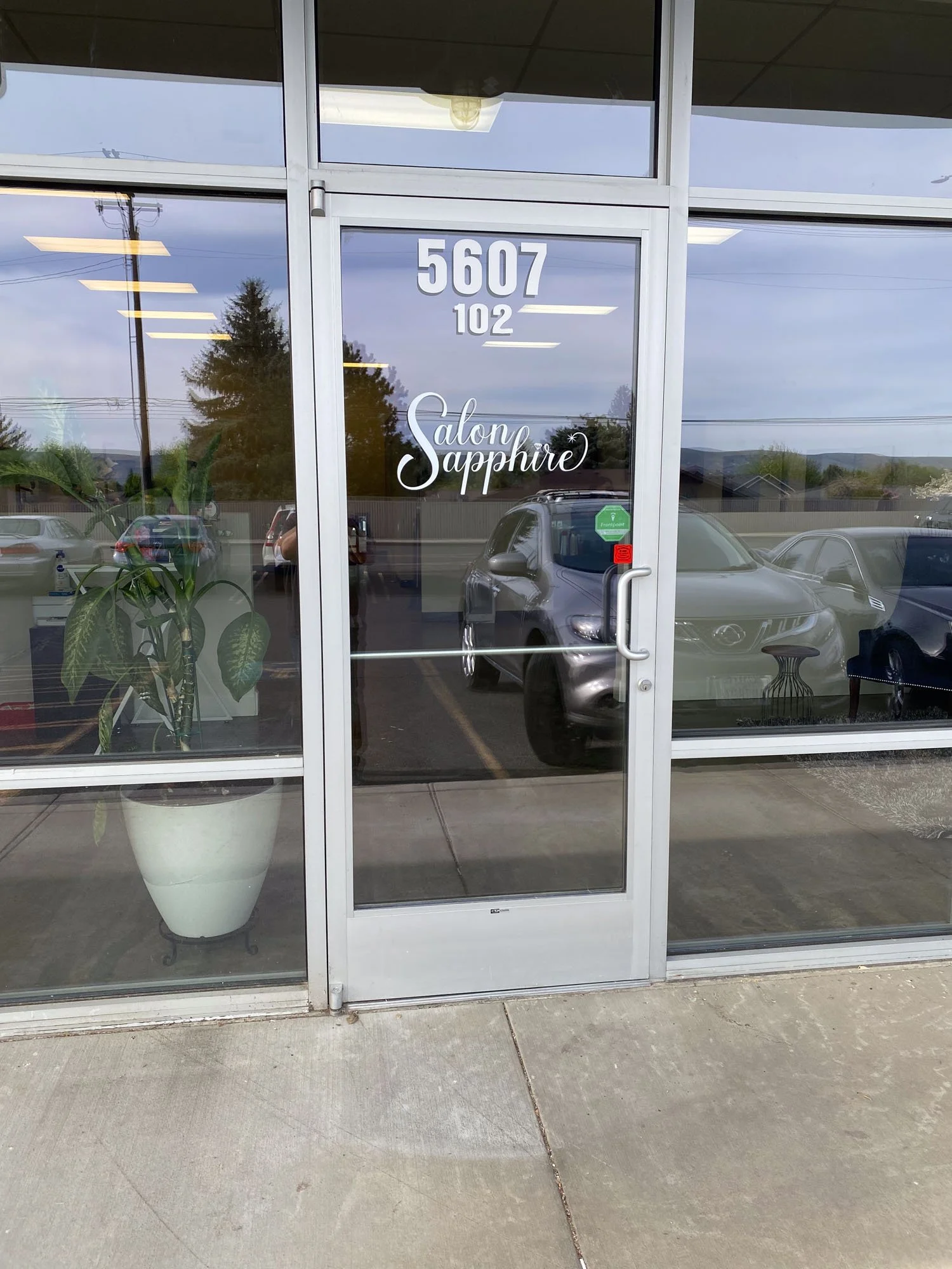 The front entrance of a beauty salon named Salon Sapphire, with large glass doors displaying the address 5607 and 102, and the salon's name in cursive lettering on the door. There are reflections of cars and a landscape visible in the glass.