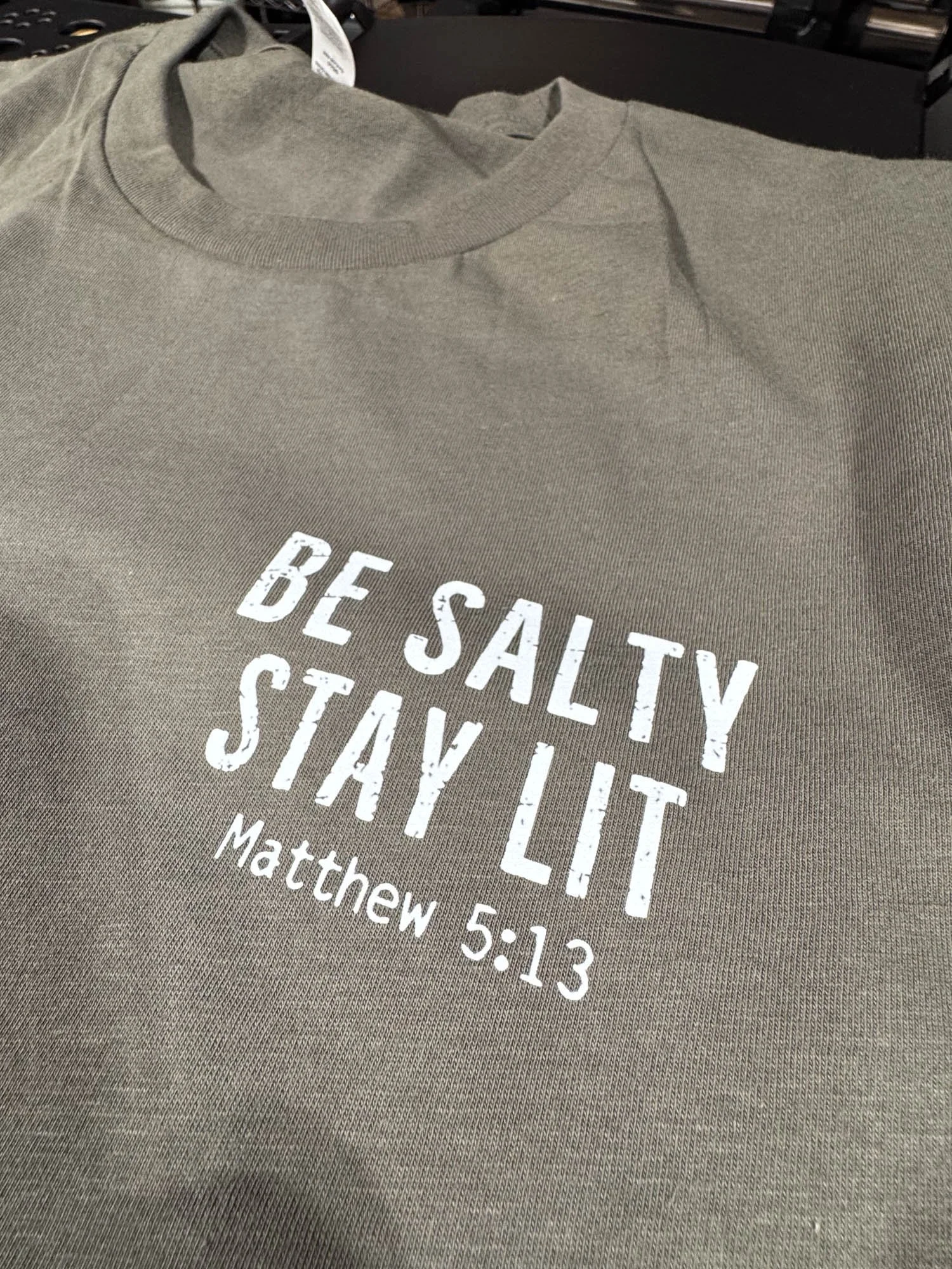 A gray T-shirt with white text that reads "Be Saltly Stay Lit" and a Bible reference Matthew 5:13.