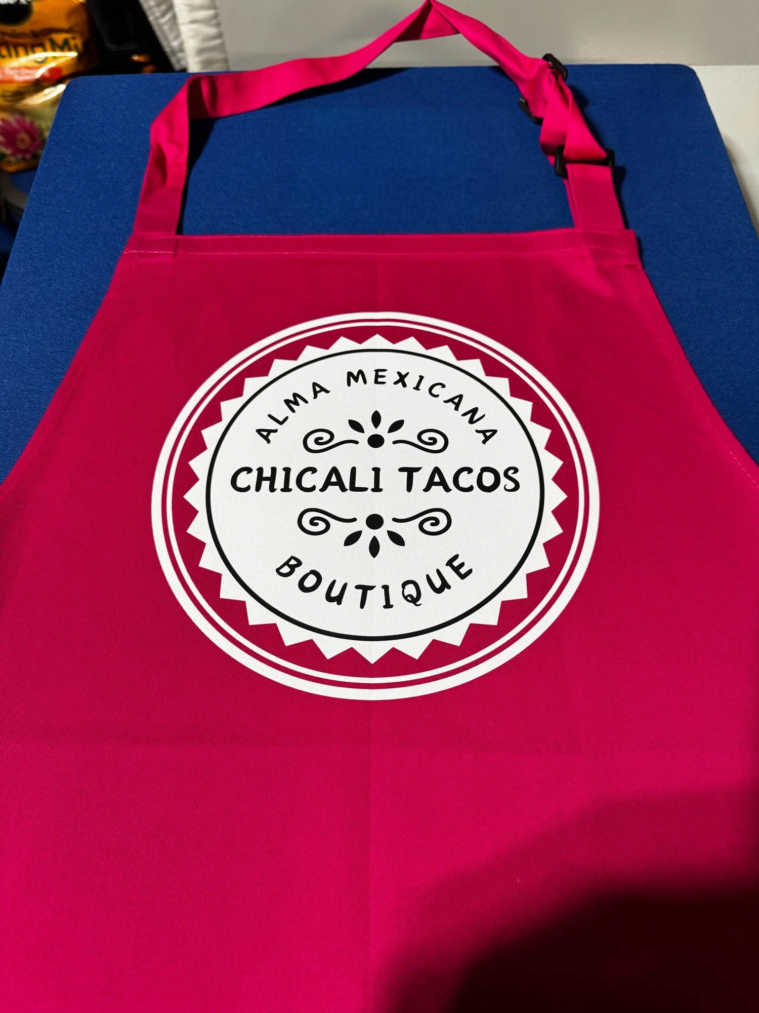 Pink reusable shopping bag with a circular black and white logo reading "Alma Mexicana Boutique Chicali Tacos" on a dark blue surface.
