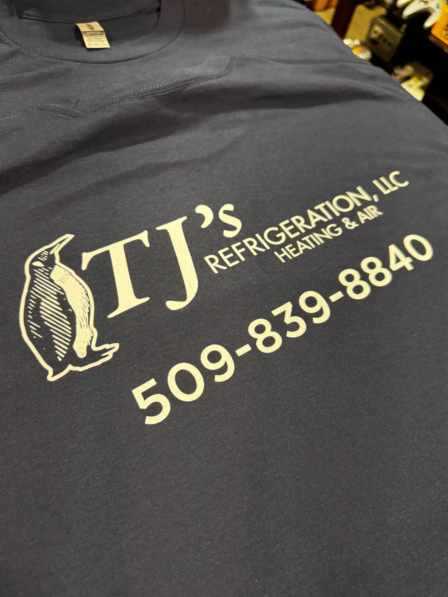 Dark gray work shirt with company logo and contact information printed on the front, featuring a penguin graphic and text for TJ's Refrigeration, LLC.