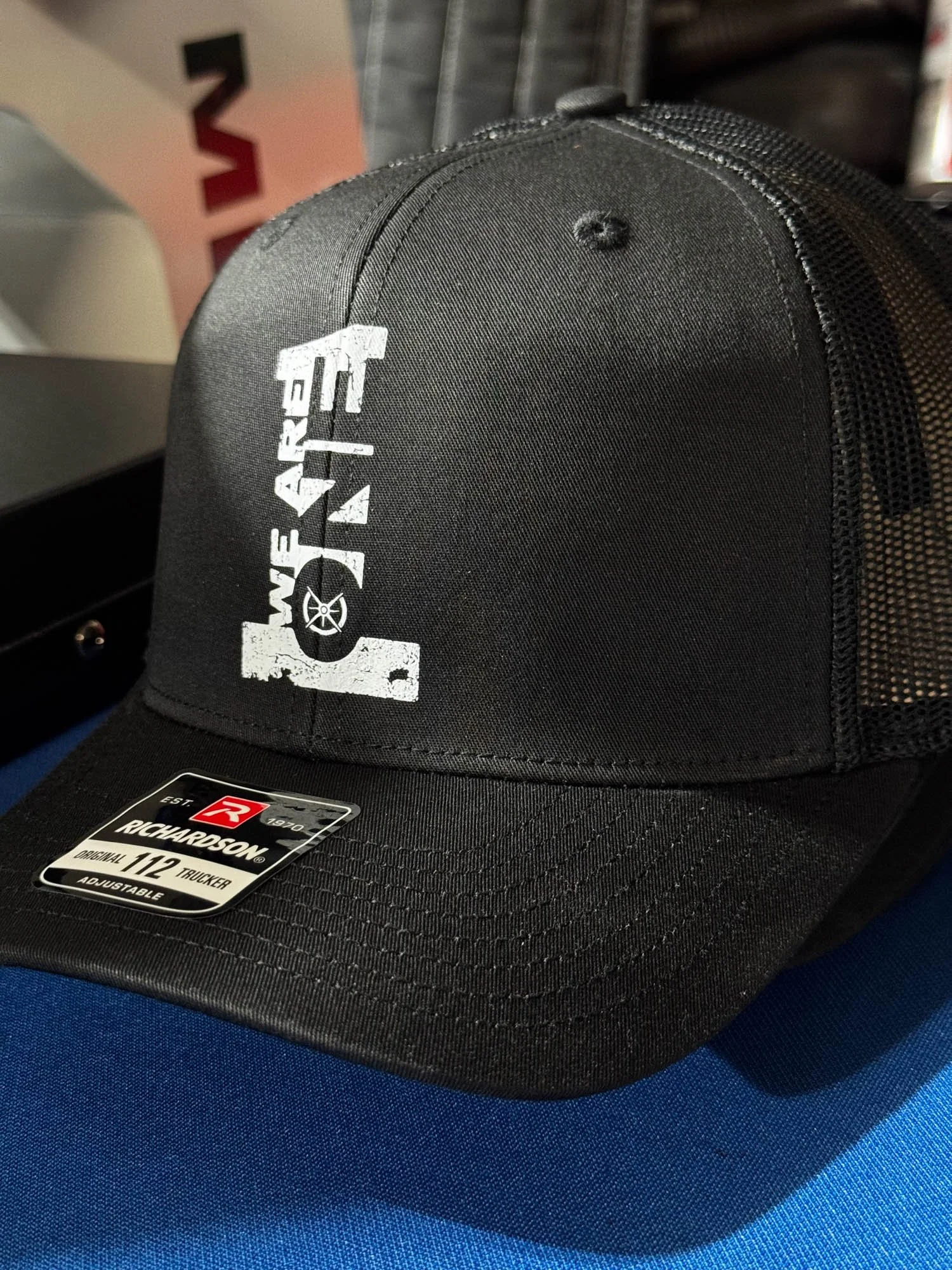 Black Richardson 112 trucker cap with white 'WE ARE TALL' logo on front, resting on a blue surface.