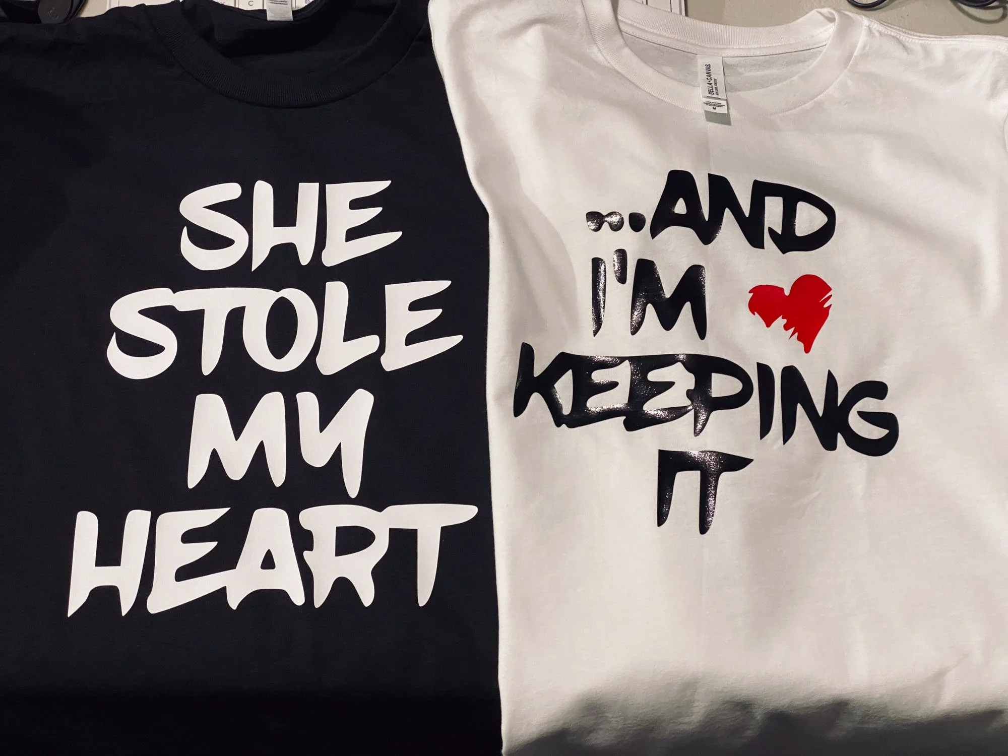 Two T-shirts hanging side by side, one black with white text and one white with black text and a red heart. The black shirt says 'SHE STOLE MY HEART.' The white shirt says 'and I’m keeping it,' with the red heart replacing the word 'heart'.