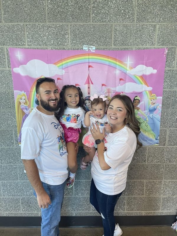 Family of four with two young girls posing in front of a colorful princess-themed backdrop featuring a rainbow, castle, and princesses, at a birthday celebration.