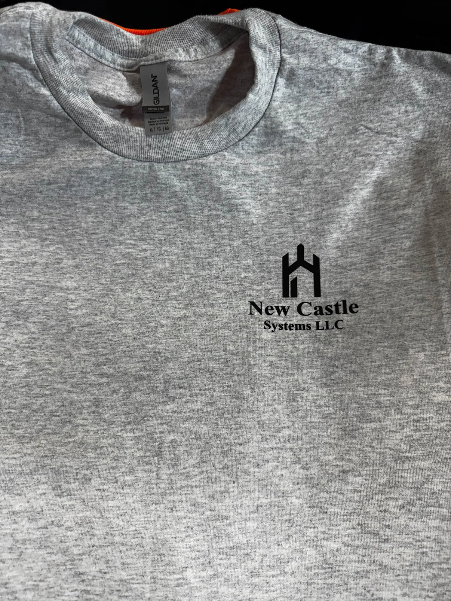 A gray T-shirt with a logo and text on the left chest area, featuring a black graphic resembling a castle tower above the words "New Castle Systems LLC."