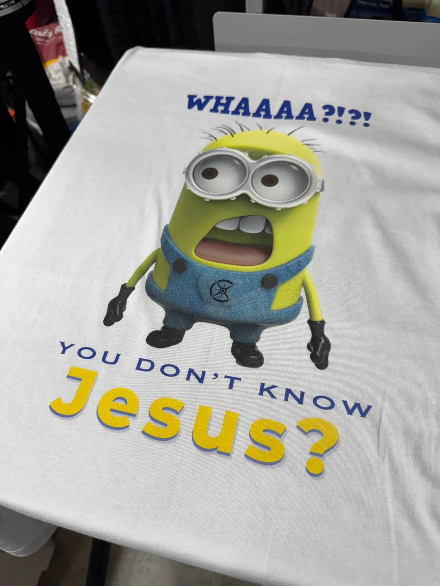 A white poster with a Minions character wearing goggles, a blue overalls, and black gloves. The Minions has an astonished expression and above it, text reads 'WHAA?!?'. Below, text reads 'You don't know Jesus?' in blue and yellow letters.