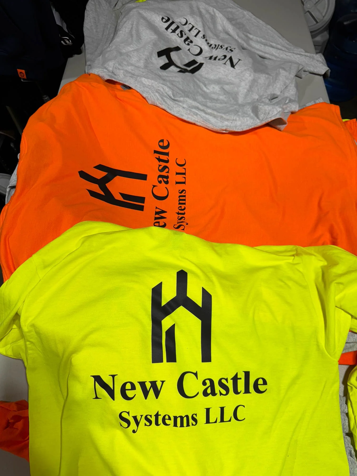 Three high-visibility safety shirts in gray, orange, and yellow, each with the logo and text for 'New Castle Systems LLC' printed on the front.