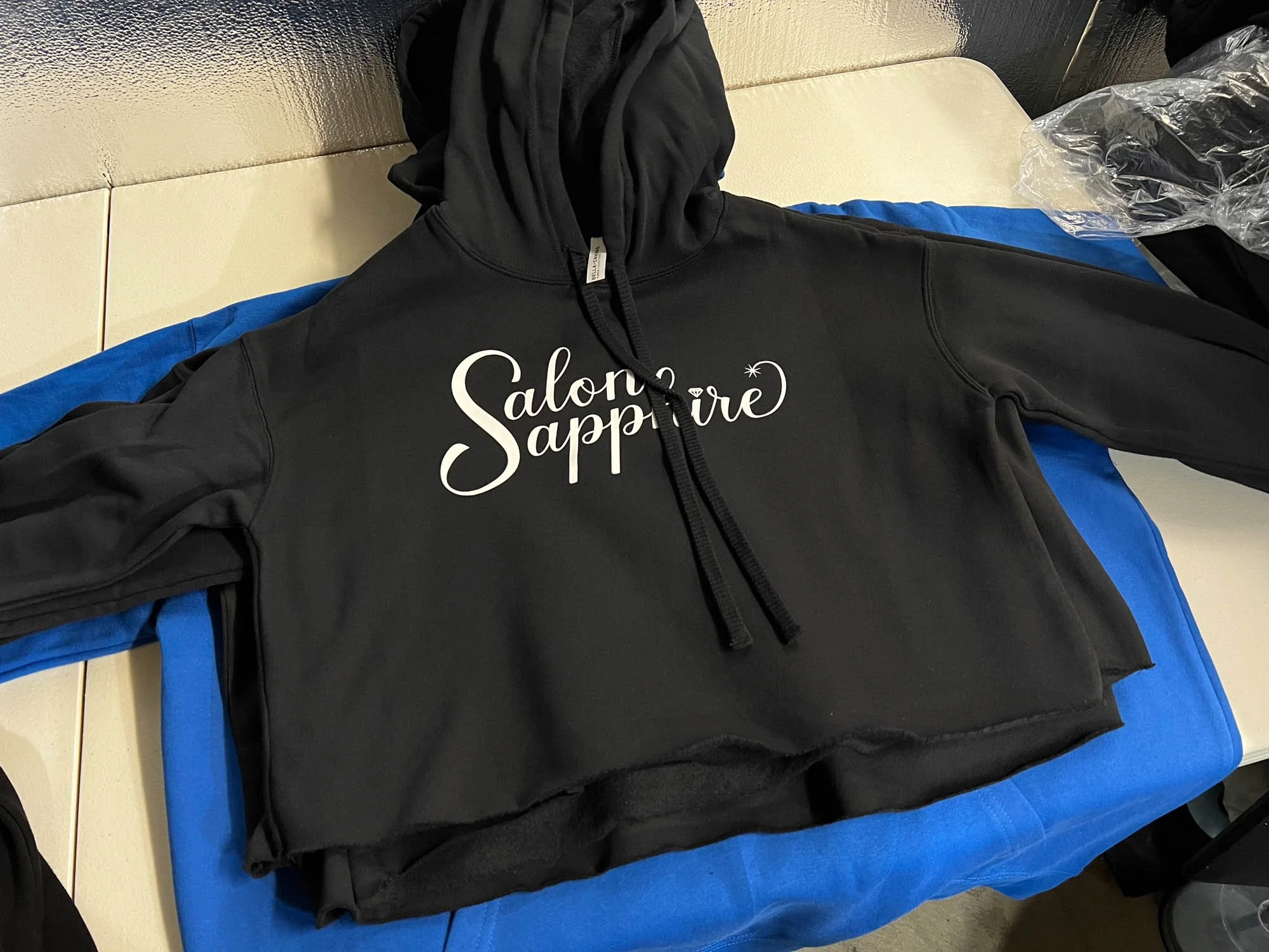 A black hoodie with white cursive text that says 'Salon Sapphira' lying on a blue surface.