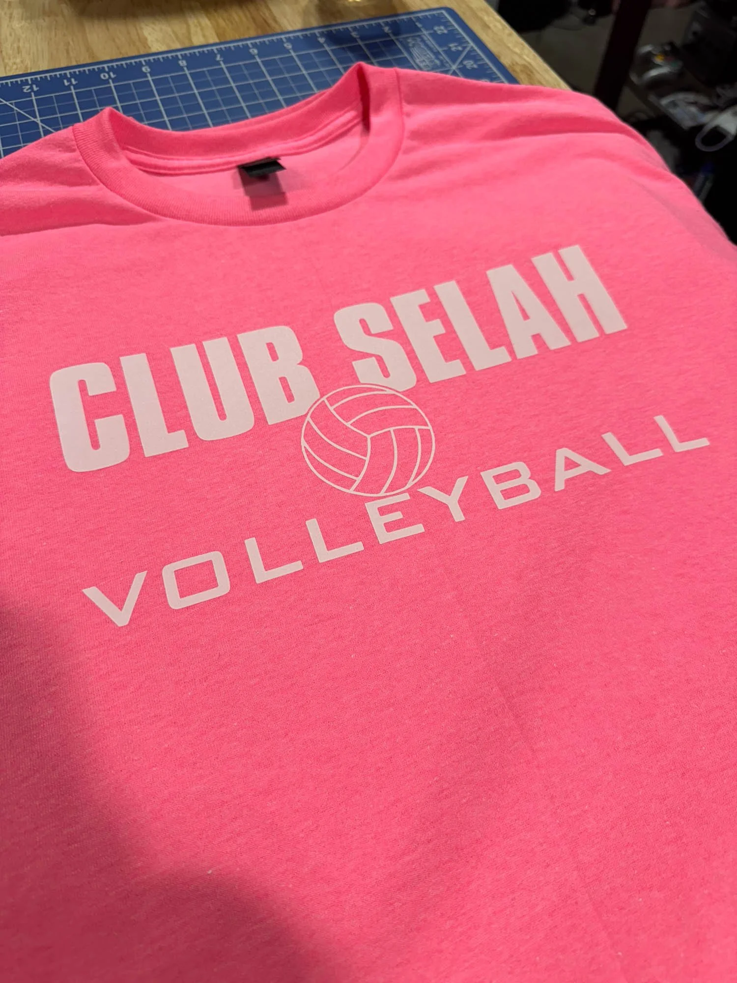 Pink T-shirt with white text that says 'CLUB SELAH VOLLEYBALL' and a volleyball graphic in the middle, lying on a cutting mat on a woodworking table.
