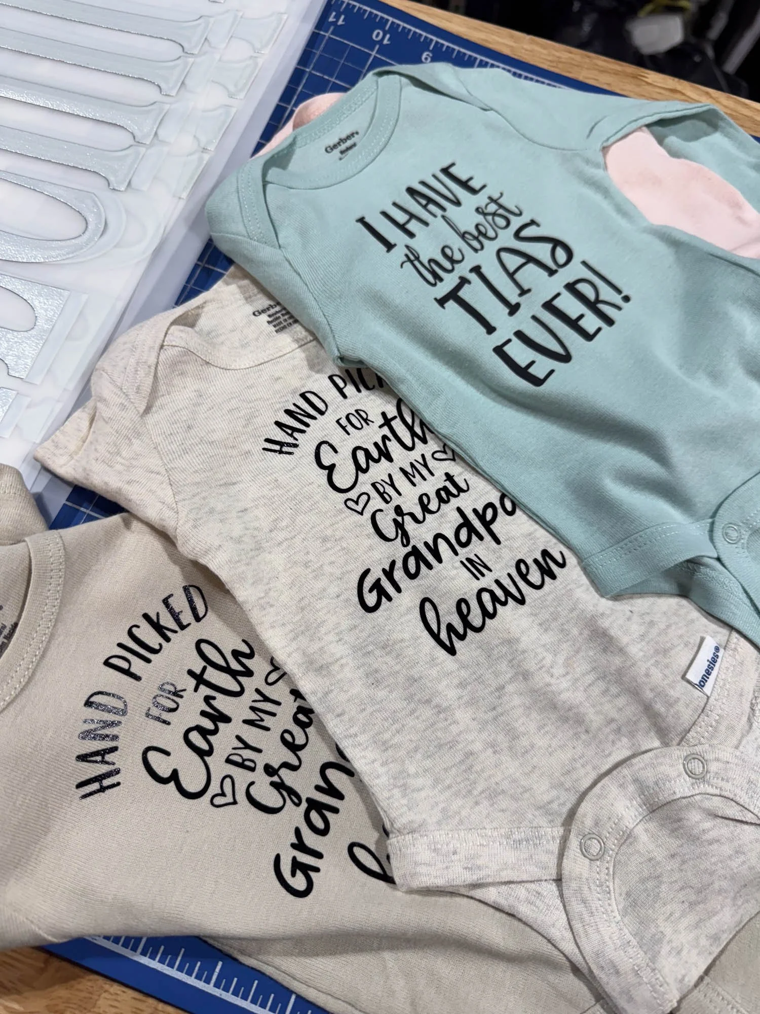 Three folded baby onesies with printed text, on a blue cutting mat. The top onesie is light blue with the words "I HAVE the best TIA EVER!" The middle onesie is gray with the words "HAND PICTURED for Earth by my Great Grandma in heaven". The bottom o