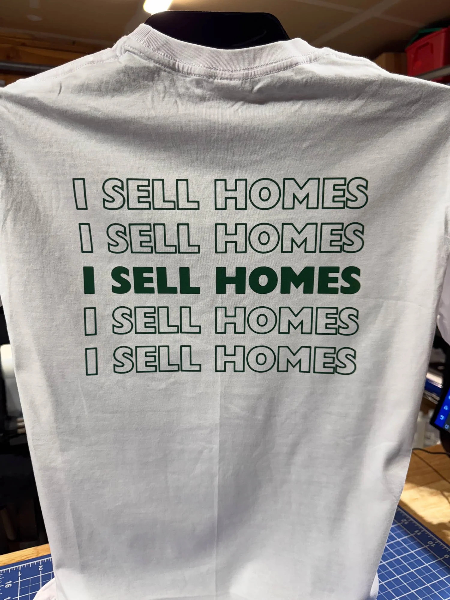 White T-shirt with the text "I SELL HOMES" repeated five times, with the third line in bold and darker green.