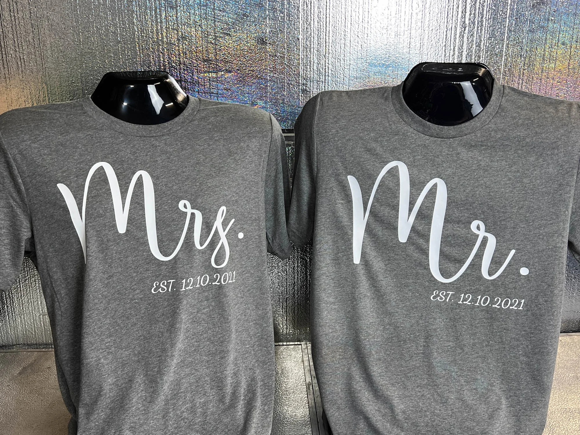 Two gray T-shirts on black mannequins with the words 'Mrs.' and 'Mr.' written in large white script, and underneath, 'EST. 12.10.2021' in smaller white text