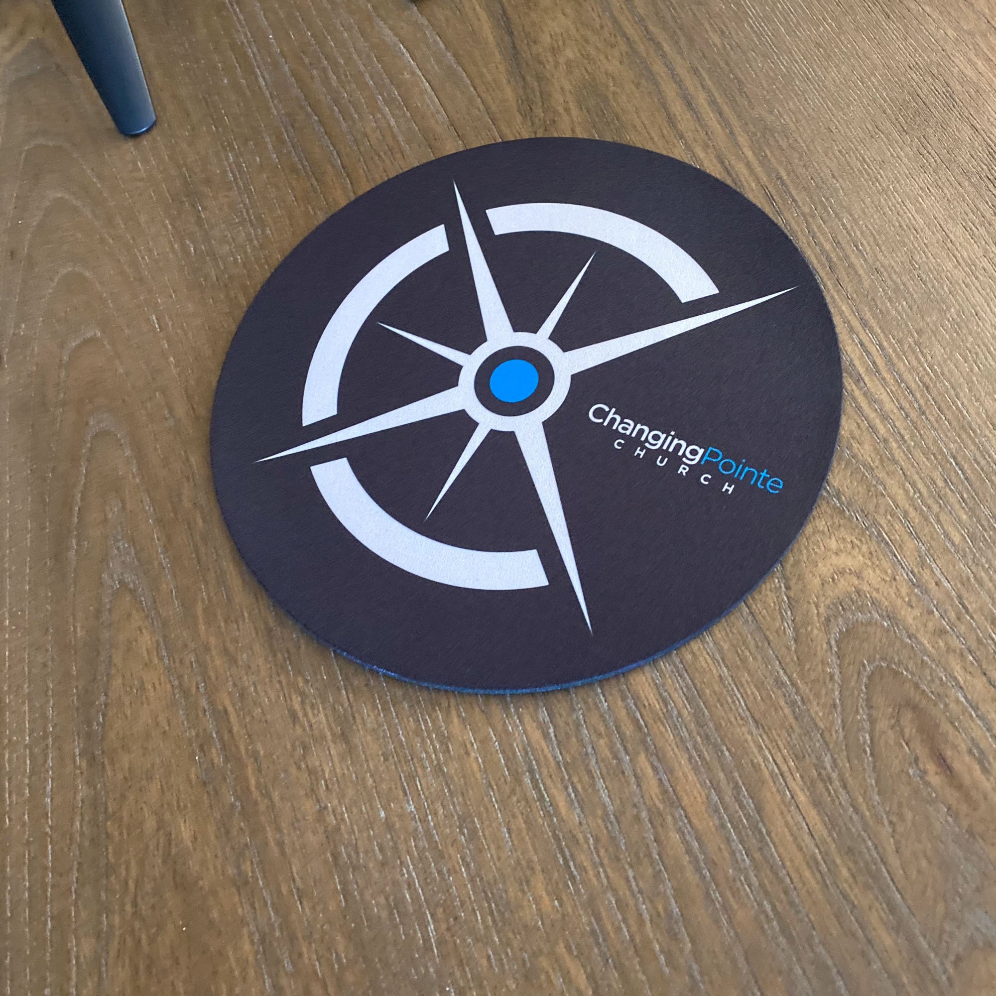 Round black mat with a white and blue compass design and the text "Changing Pointe Church" on a wooden floor.