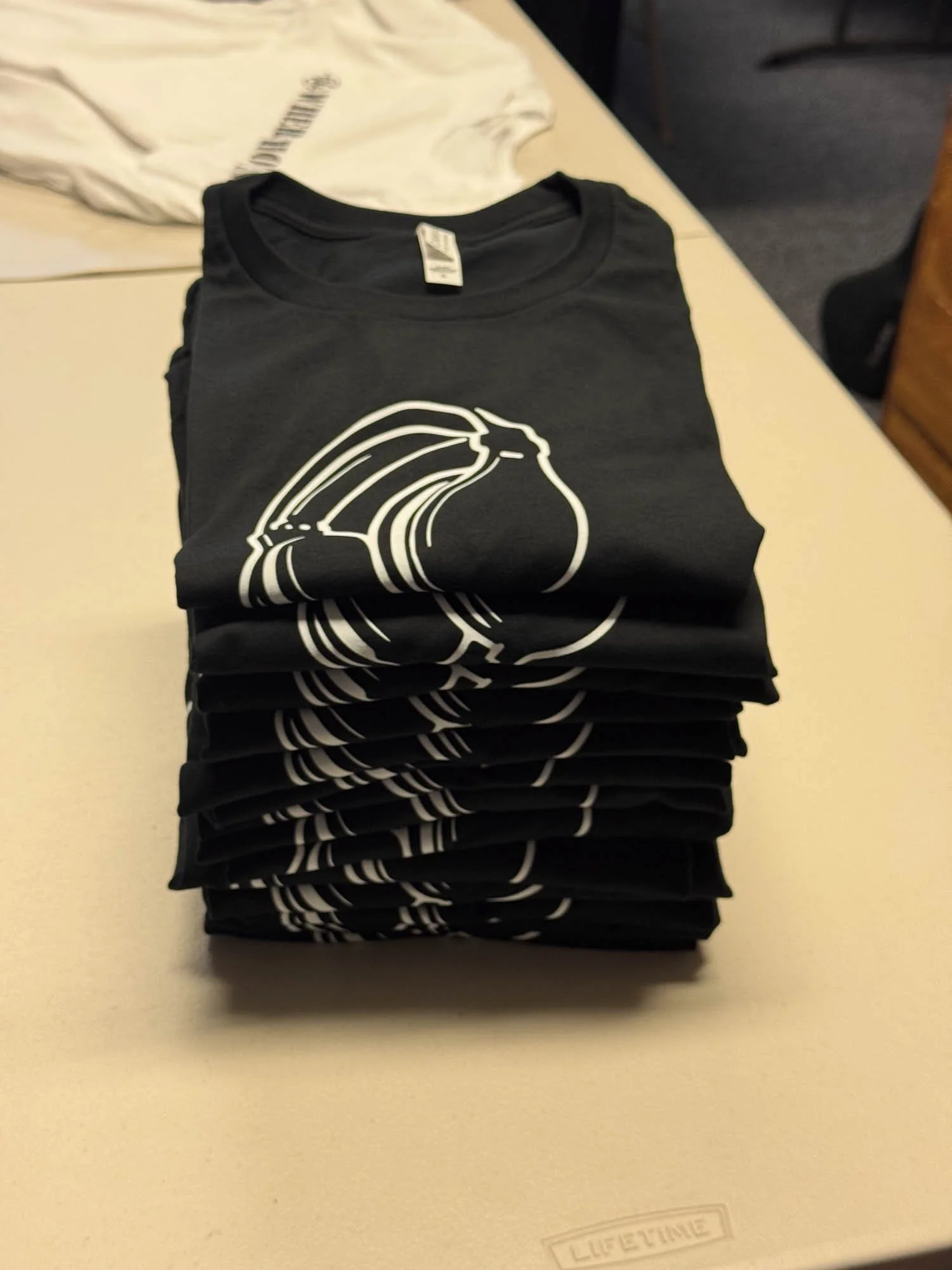 A stacked pile of black t-shirts with a white line drawing of a pear on the front, placed on a beige surface.